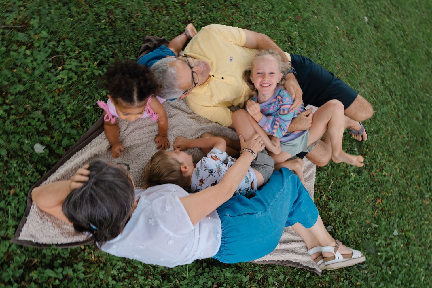 Family laughing and cuddling together on a blanket in green grass during an outdoor photo session.