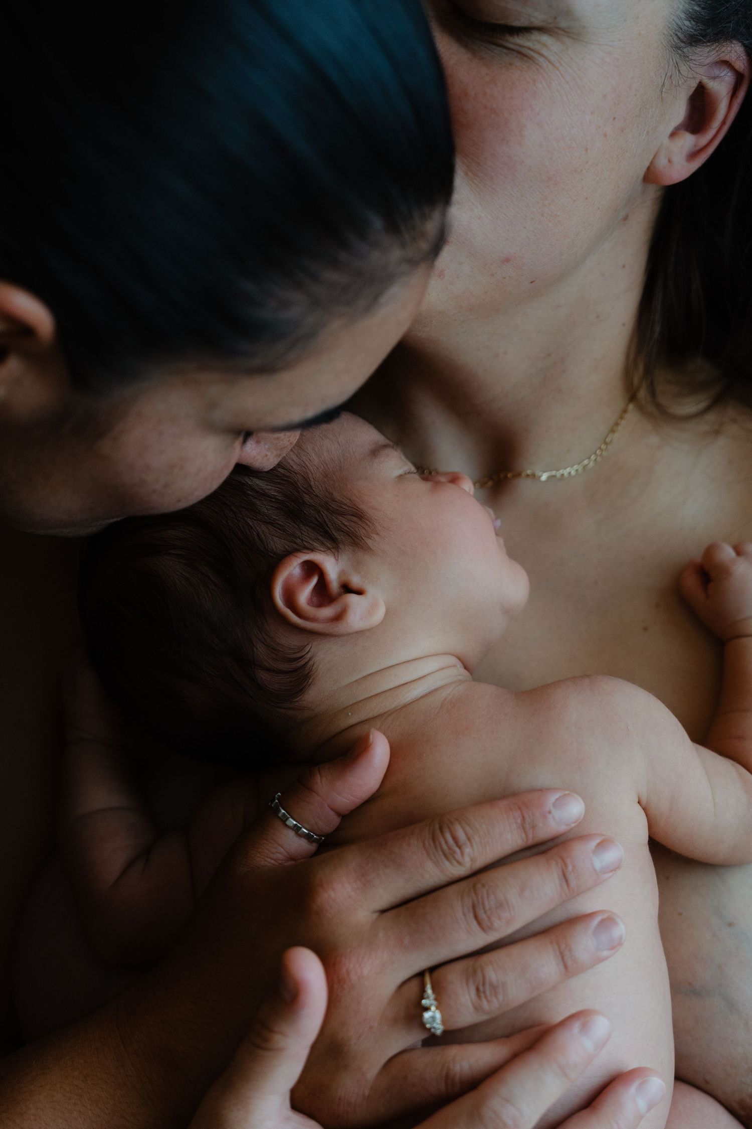 Two mothers tenderly kissing their newborn baby, captured in an intimate and emotional portrait.