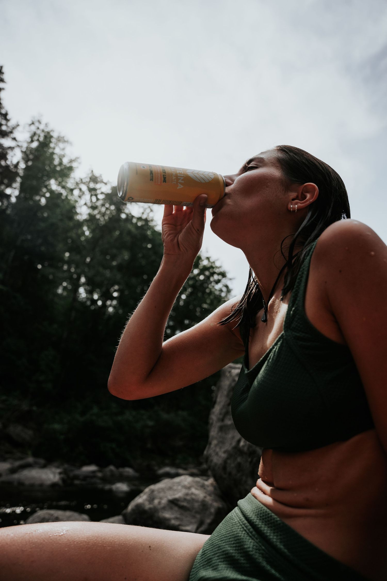 Young woman in green bikini drinking from a bottle while sitting on rocks outdoors.