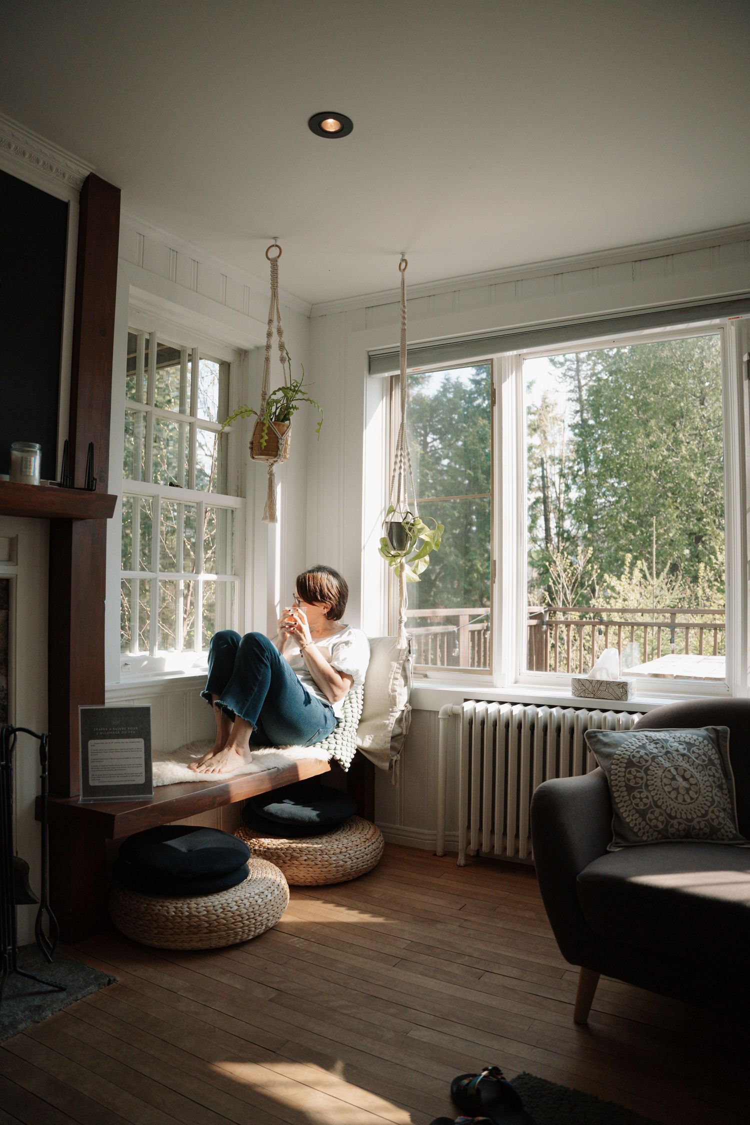 Woman relaxing in hanging macrame chair by large windows in cozy sunlit living room with houseplants.