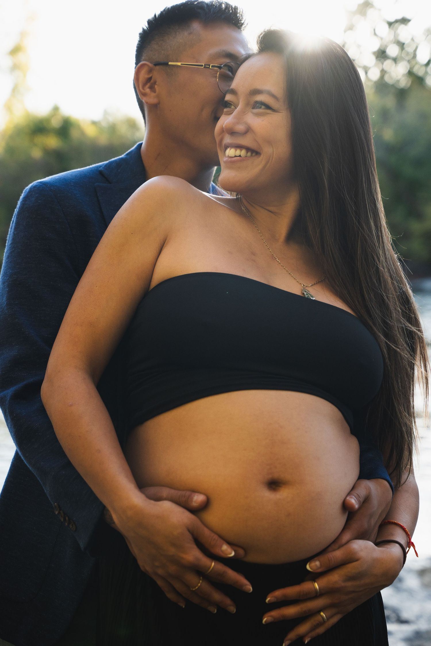 Couple embracing outdoors at sunset, pregnant woman smiling as partner kisses her cheek while cradling her baby bump.