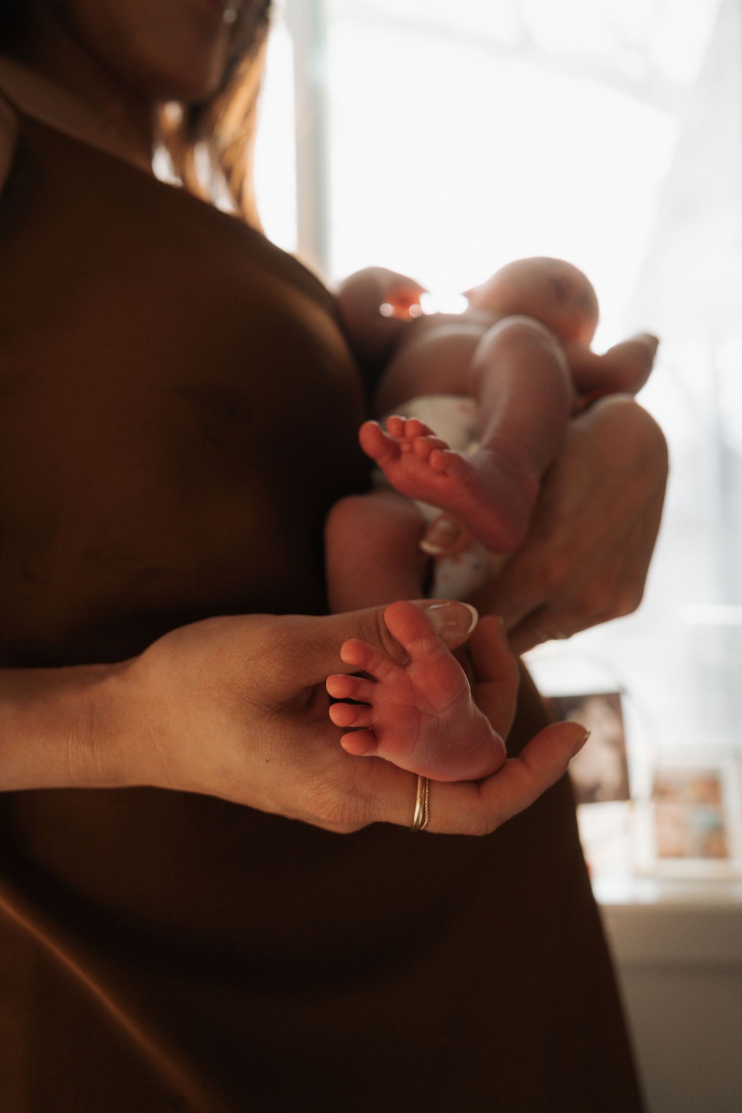 Mother holding newborn baby while another hand gently cradles the infants tiny feet in warm light.