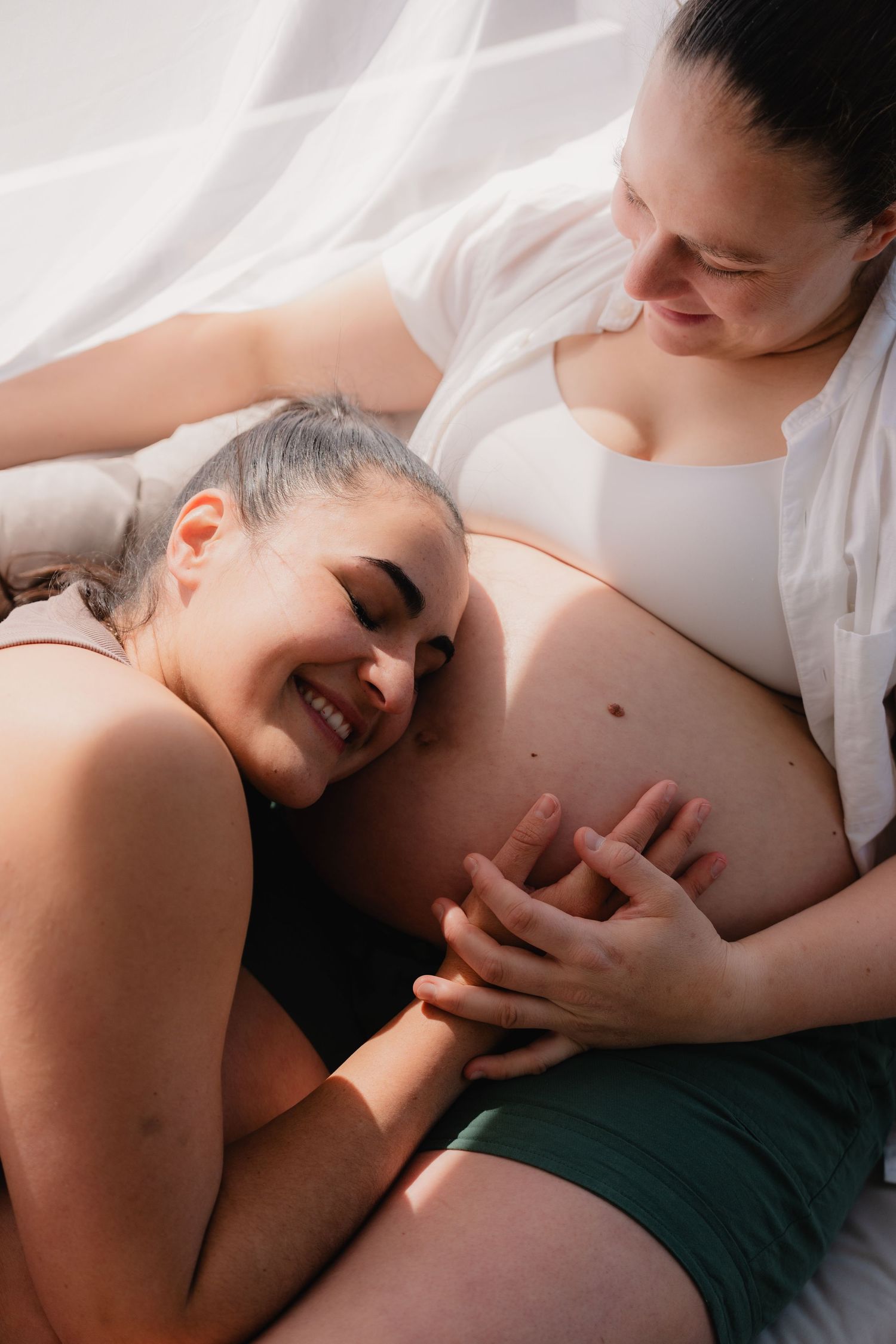 A smiling woman resting her head on her pregnant partners belly while holding hands in soft natural light.