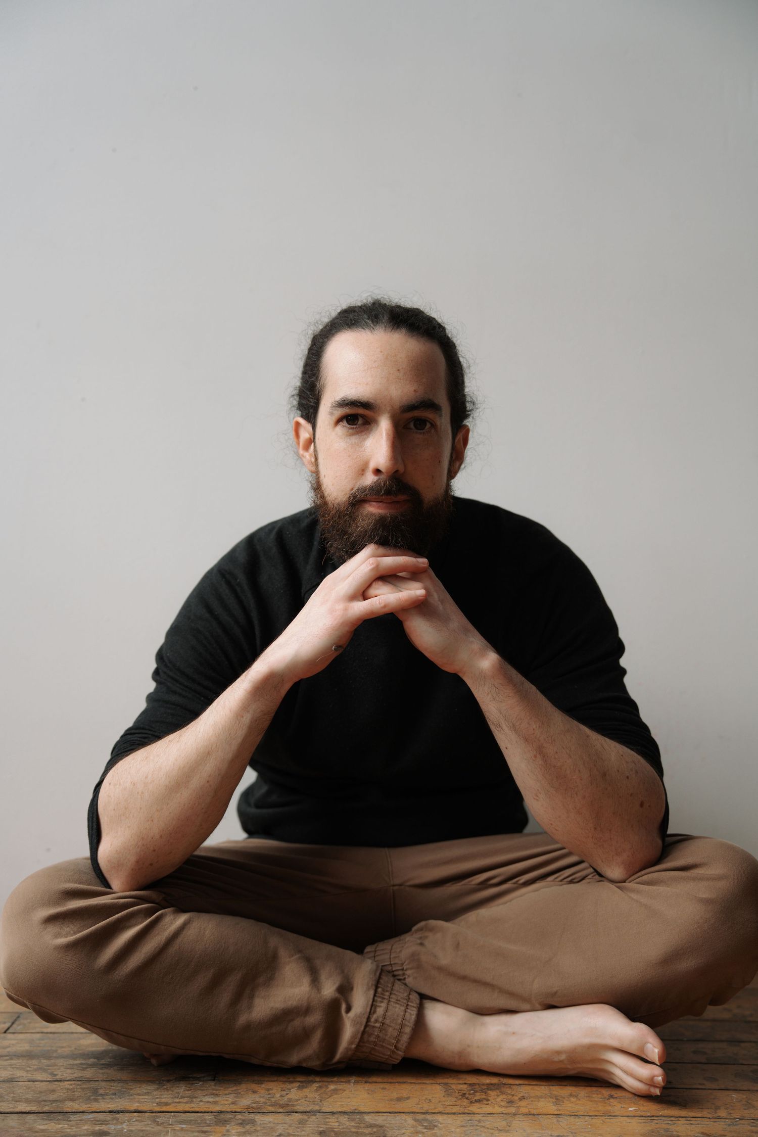 Bearded man with tied back hair sitting cross-legged on wooden floor in black shirt and khaki pants.