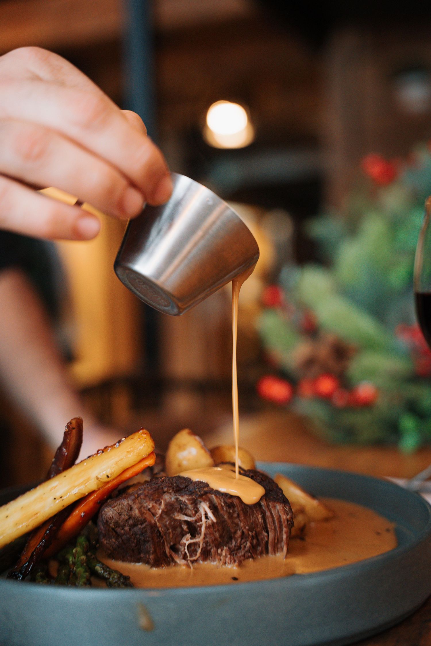 Hand pouring rich gravy from steel pitcher over braised beef with roasted vegetables on rustic plate.