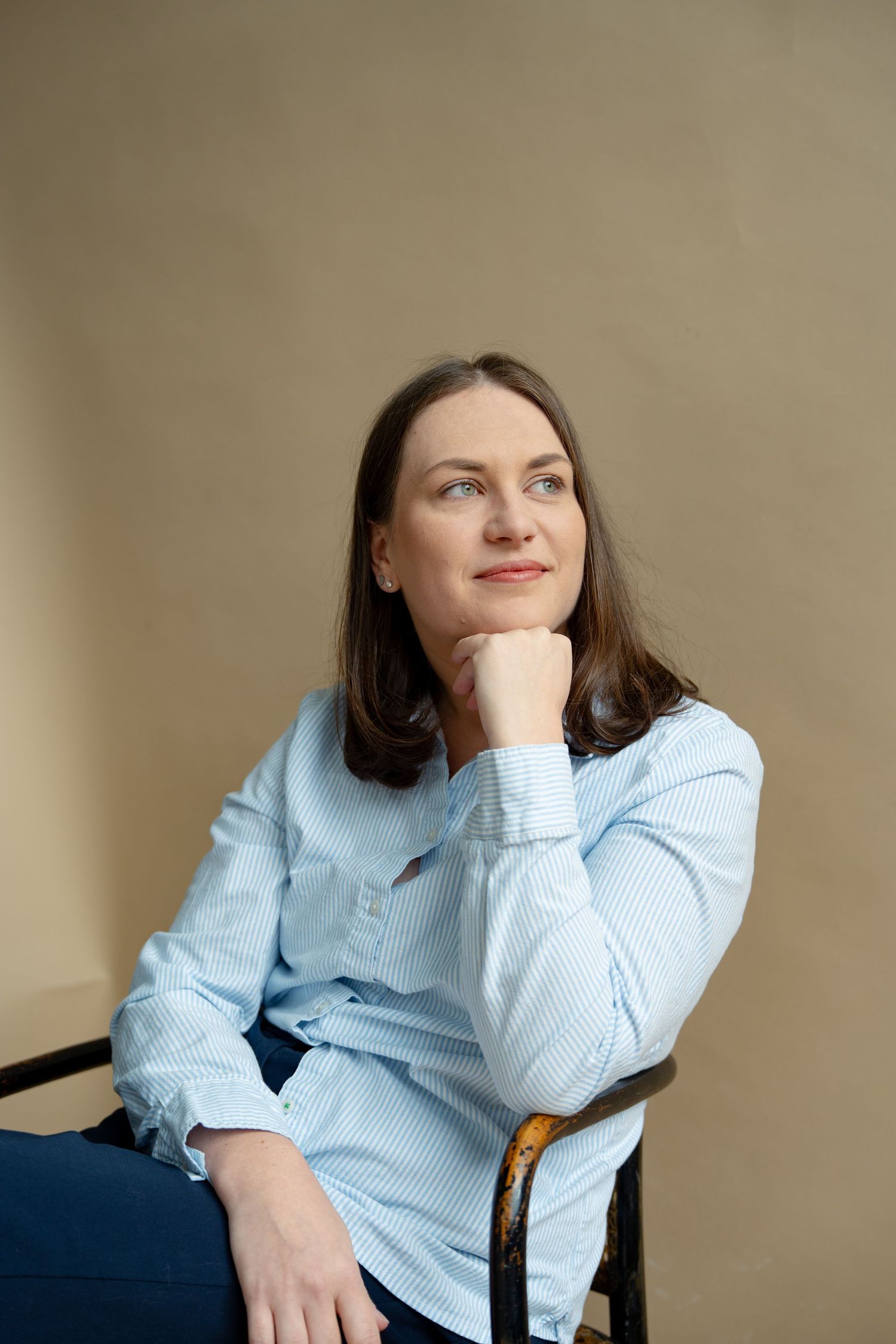 Woman in light blue striped shirt sitting on chair with hand on chin looking up.