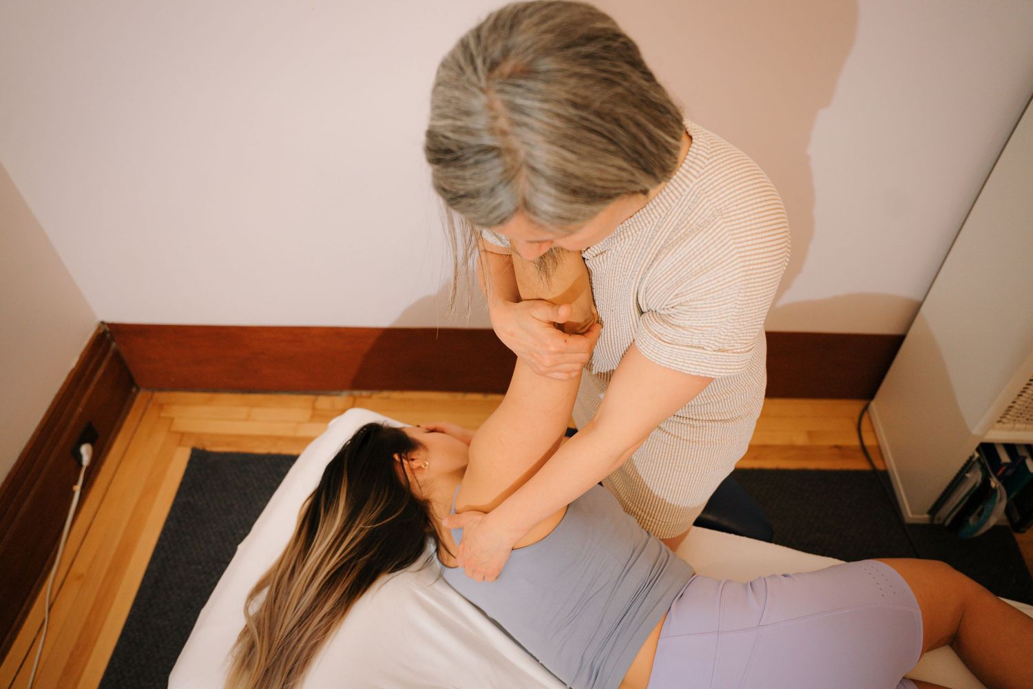 A gray-haired therapist performs a neck and shoulder massage on a client lying on a floor mat.