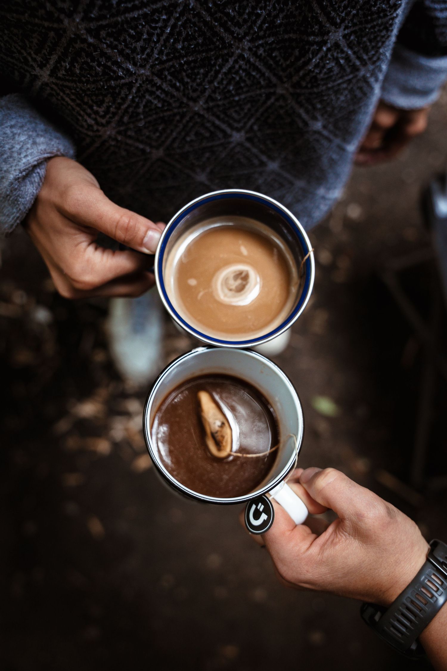 Two hands holding enamel camping mugs filled with hot coffee outdoors in nature.