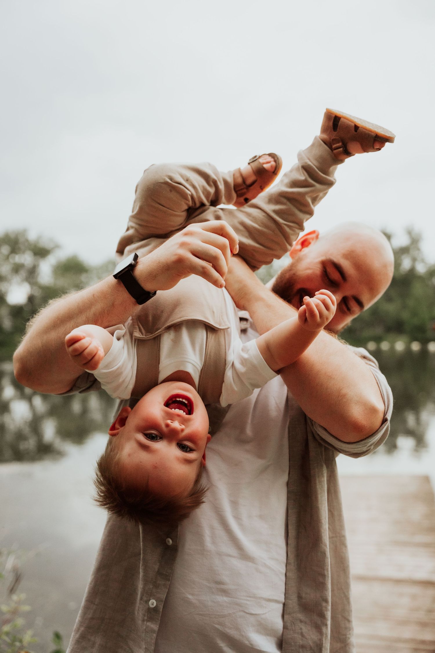 Laughing bald father playfully holds giggling toddler upside down outdoors near a lake.