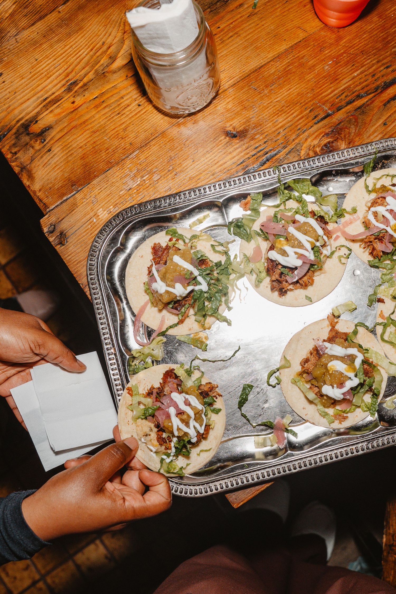 Overhead view of hands reaching for loaded tacos on silver tray with sour cream and fresh toppings on wooden table.