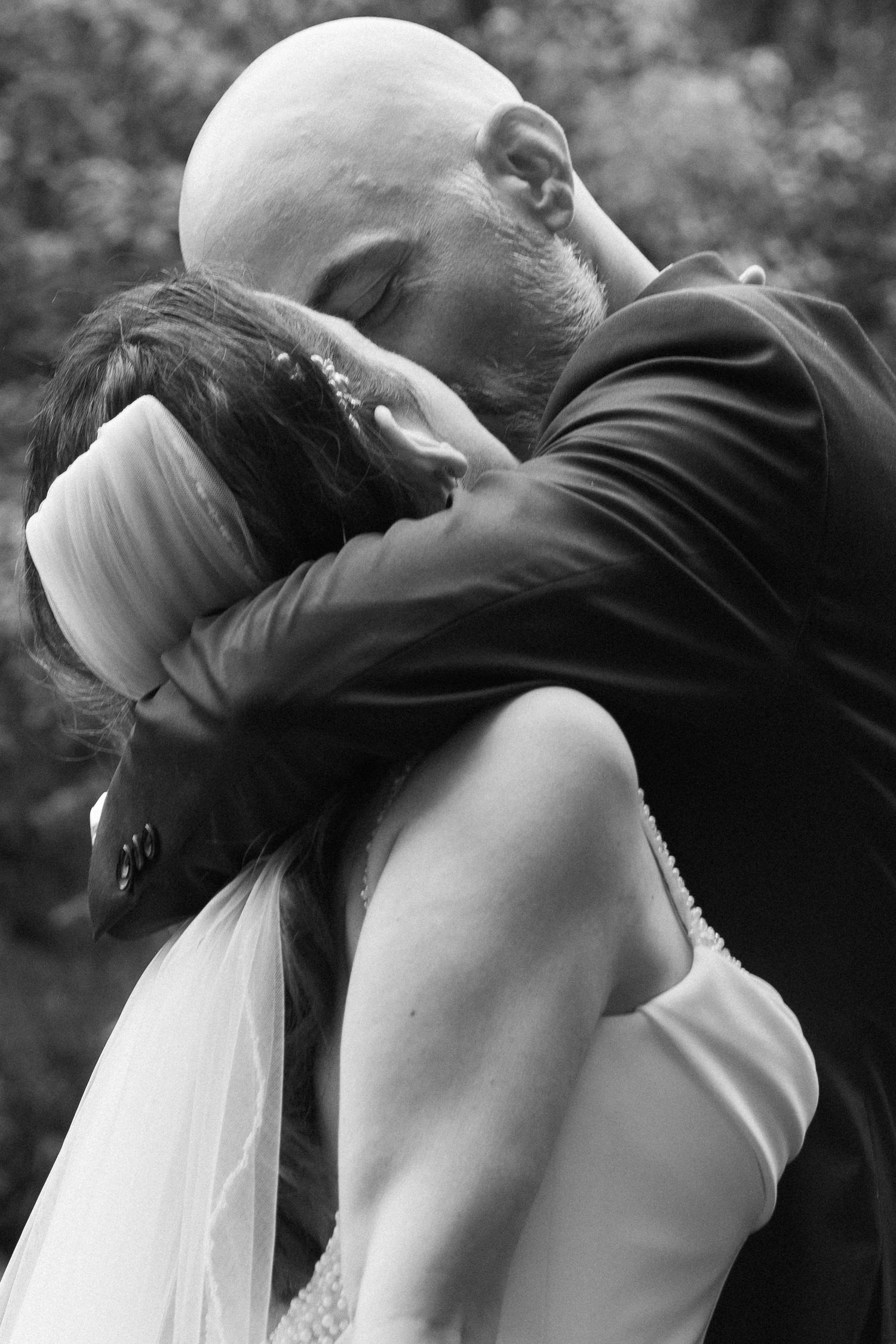 Black and white photo of a bald groom embracing his bride in a wedding veil during an emotional moment.