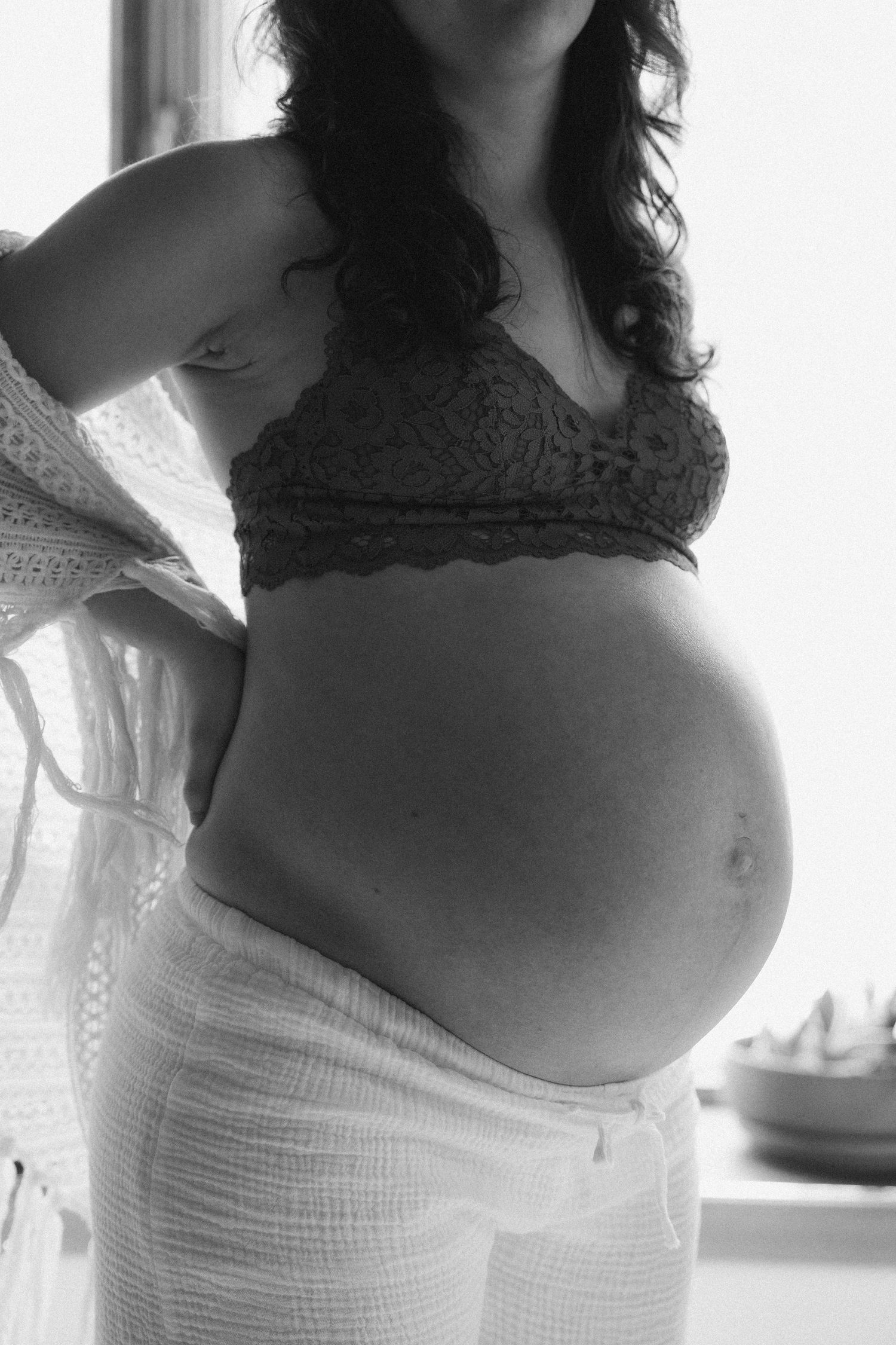 Black and white photo of a pregnant woman in a lace bralette cradling her bare baby bump indoors.