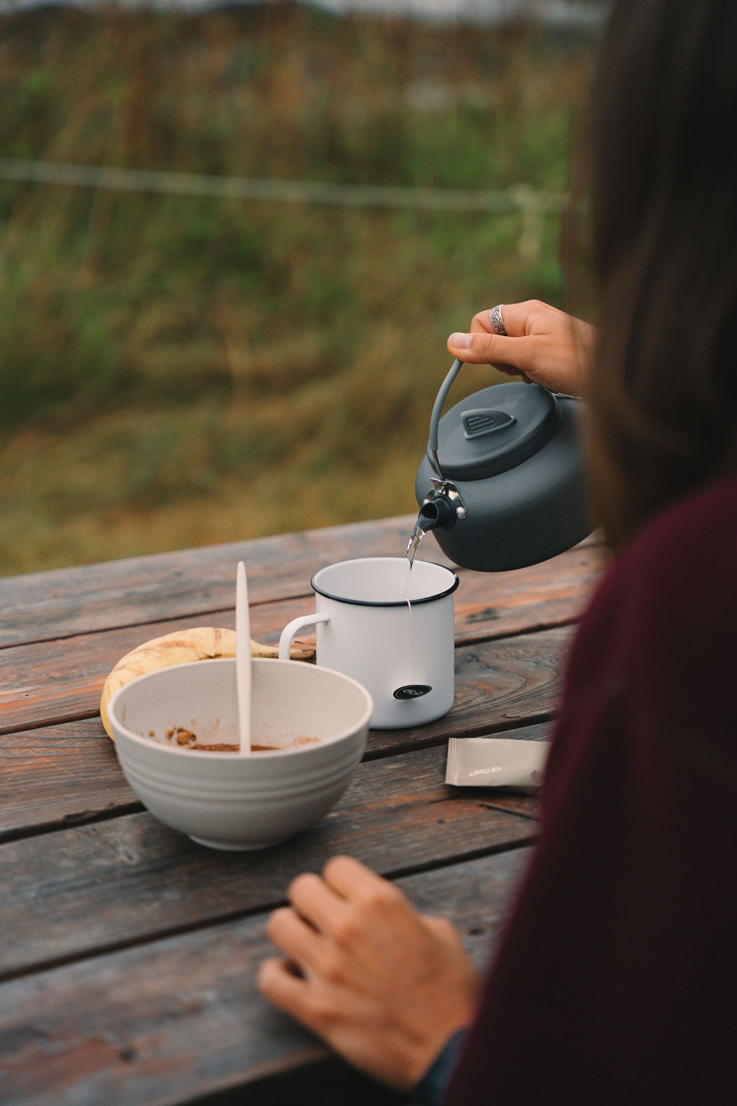 Woman pouring hot water from a camping kettle into a mug on a rustic wooden table outdoors.