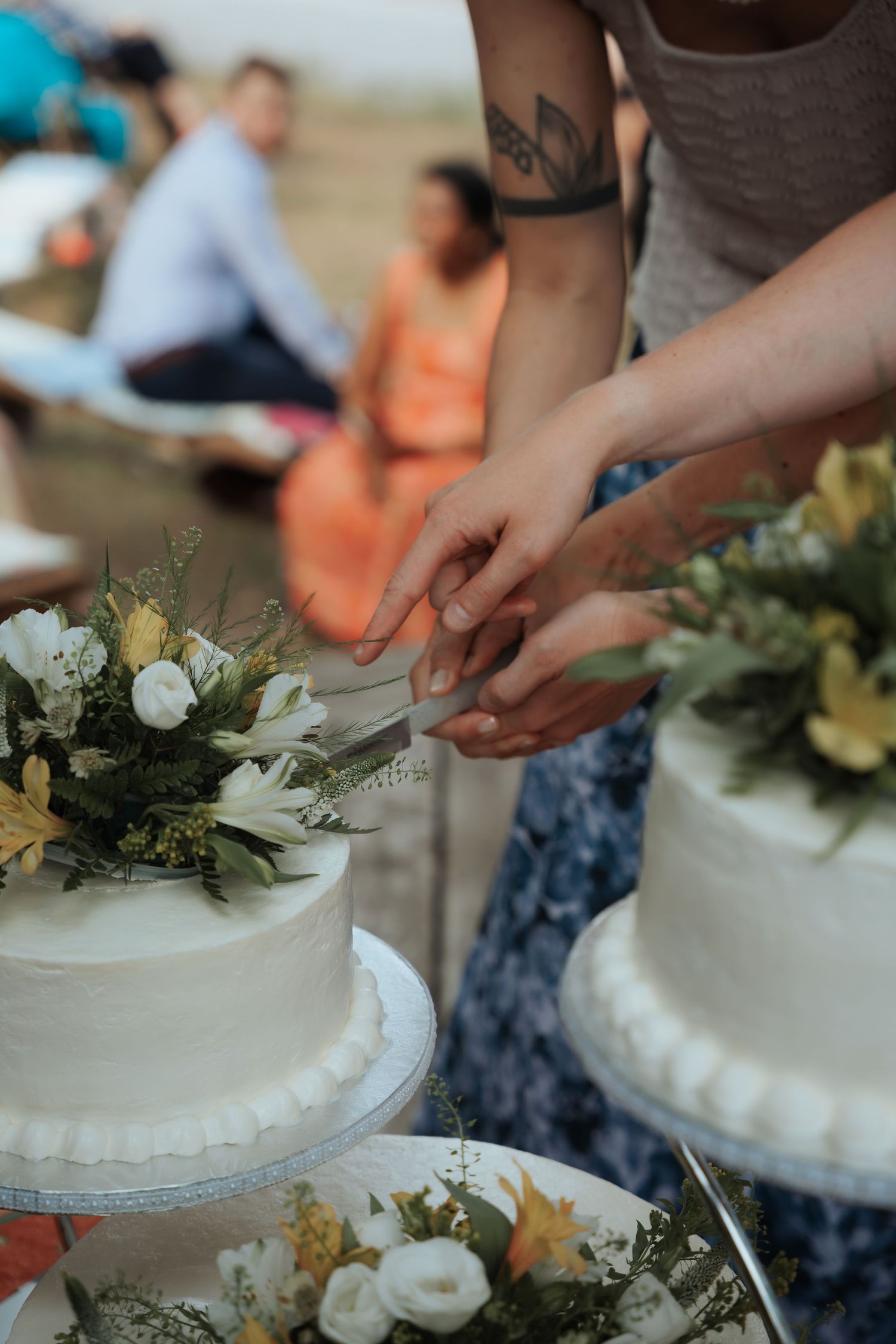 Two people with tattooed arms cut a white wedding cake decorated with yellow and white flowers outdoors.