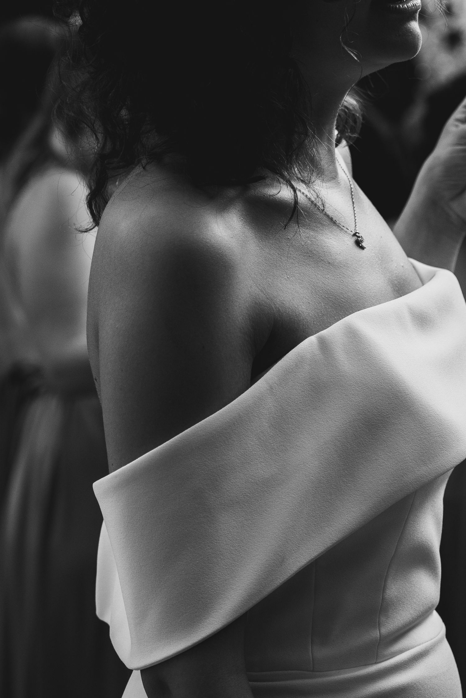 Bride in elegant off-shoulder white wedding gown with delicate necklace, black and white photo.