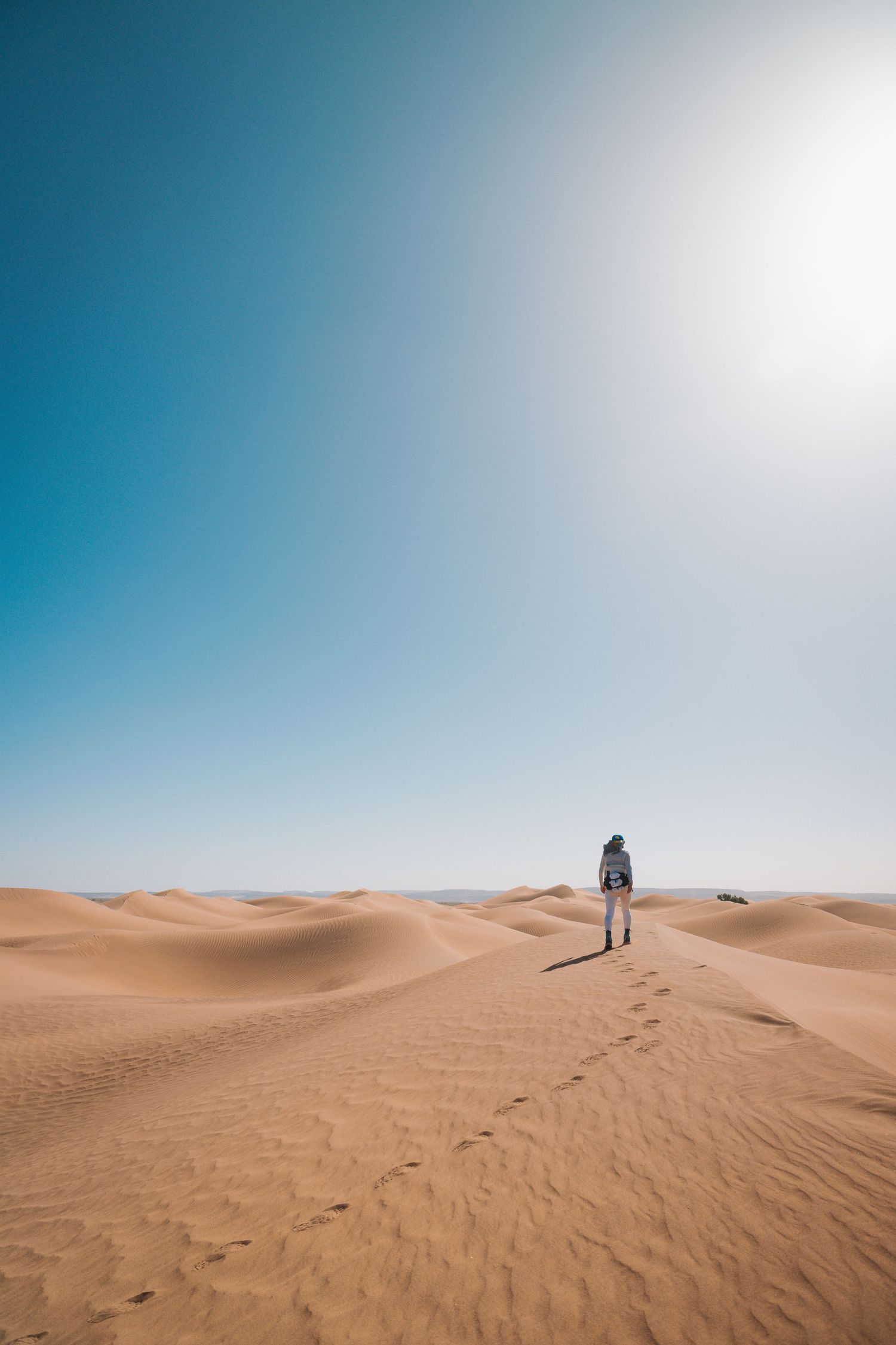 A lone hiker with a backpack stands on golden sand dunes under a bright sun in a vast desert landscape.