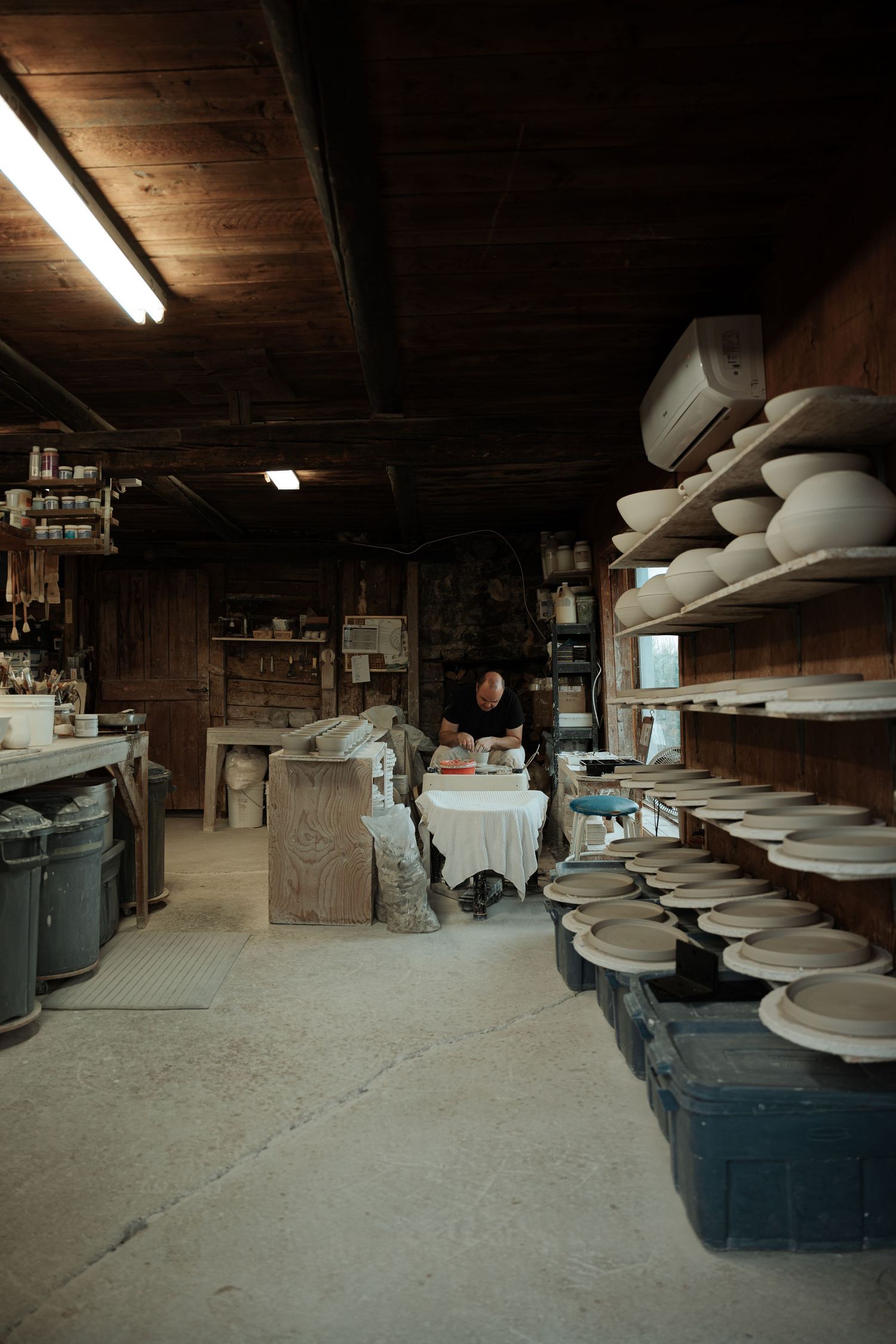Potter working in a rustic wood-paneled studio with shelves lined with ceramic bowls and plates.