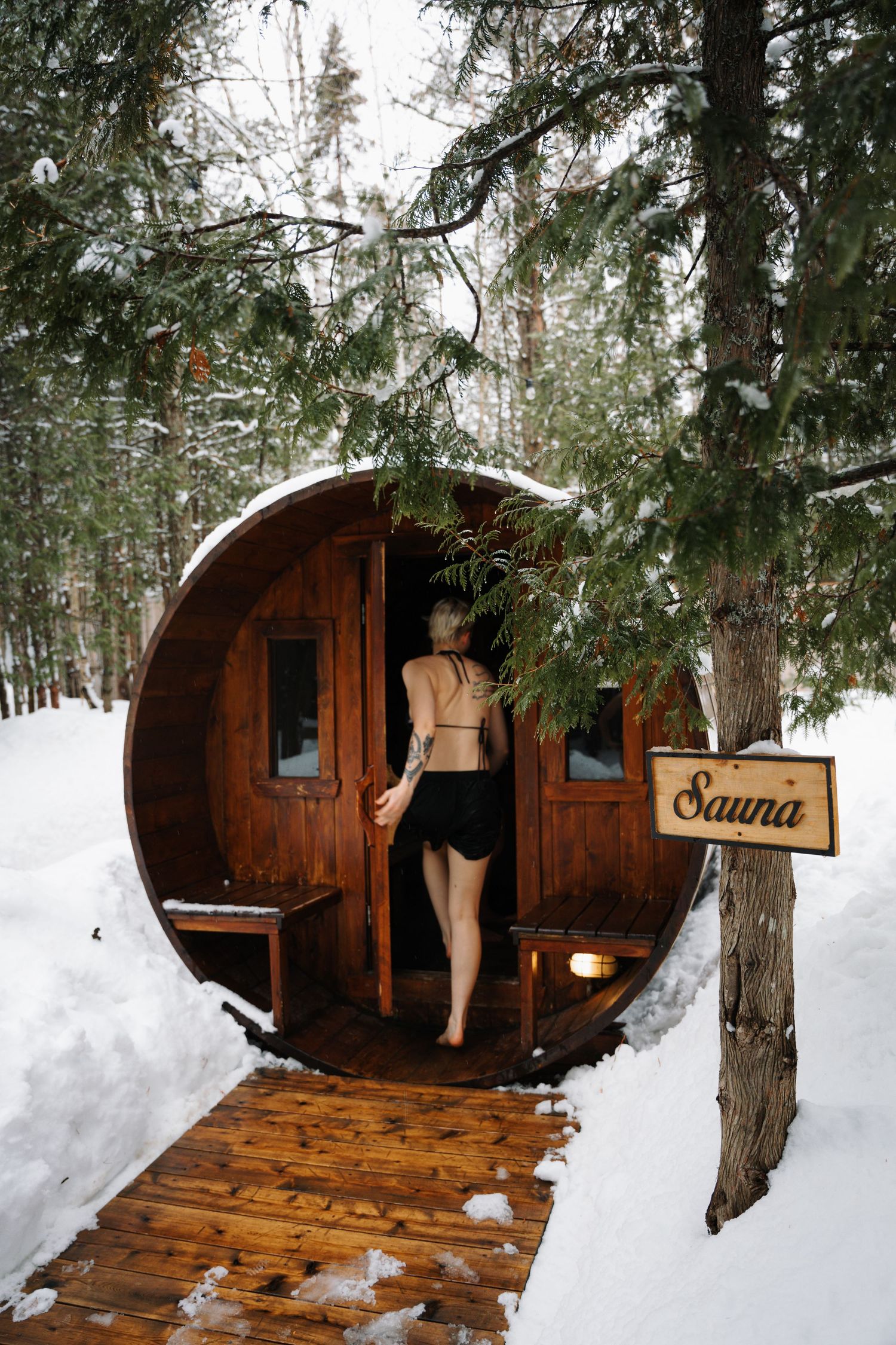 Woman in swimwear entering a barrel sauna surrounded by snow-covered pine trees in winter.