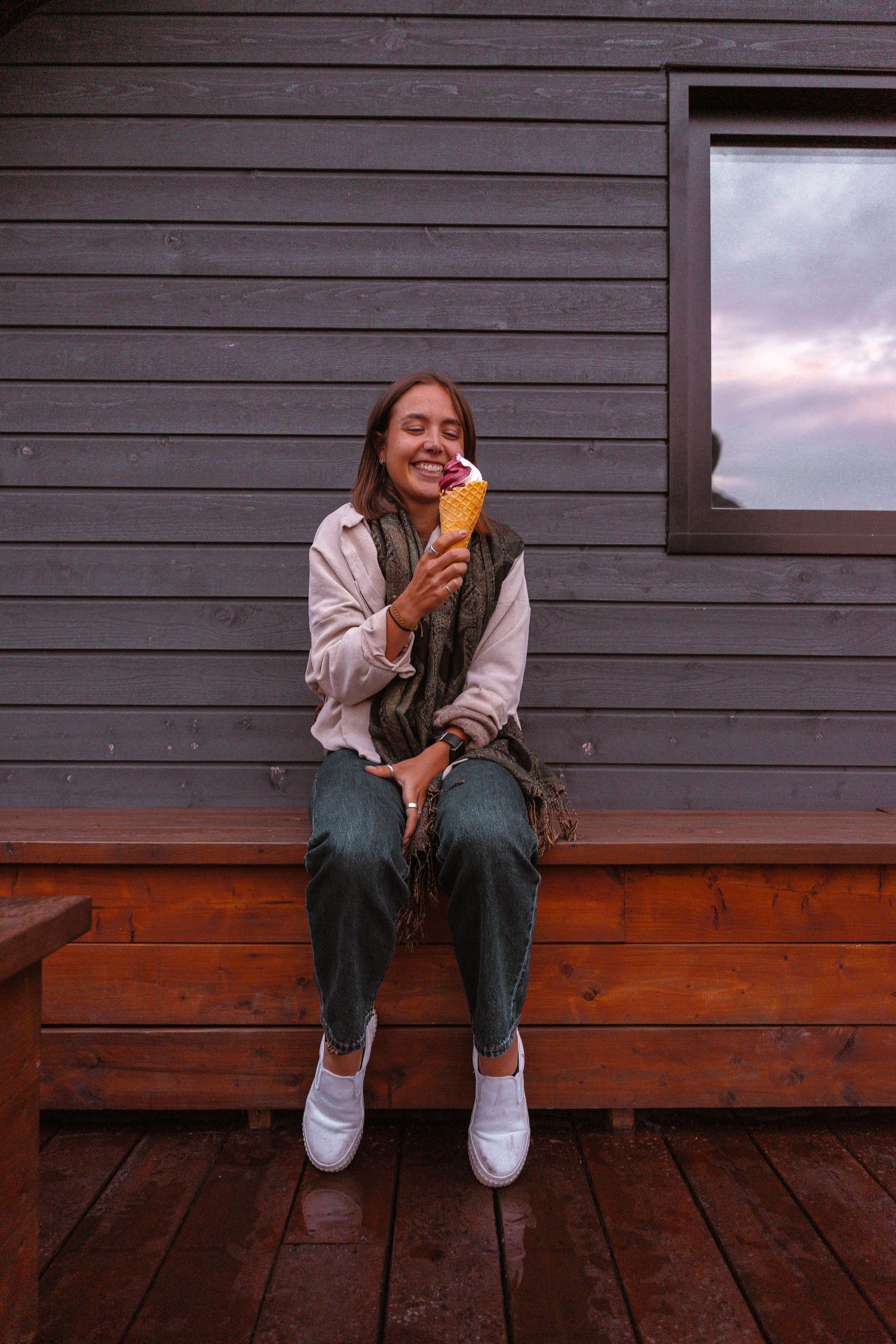 Happy woman sitting on a wooden porch eating an ice cream cone outside a gray cabin.