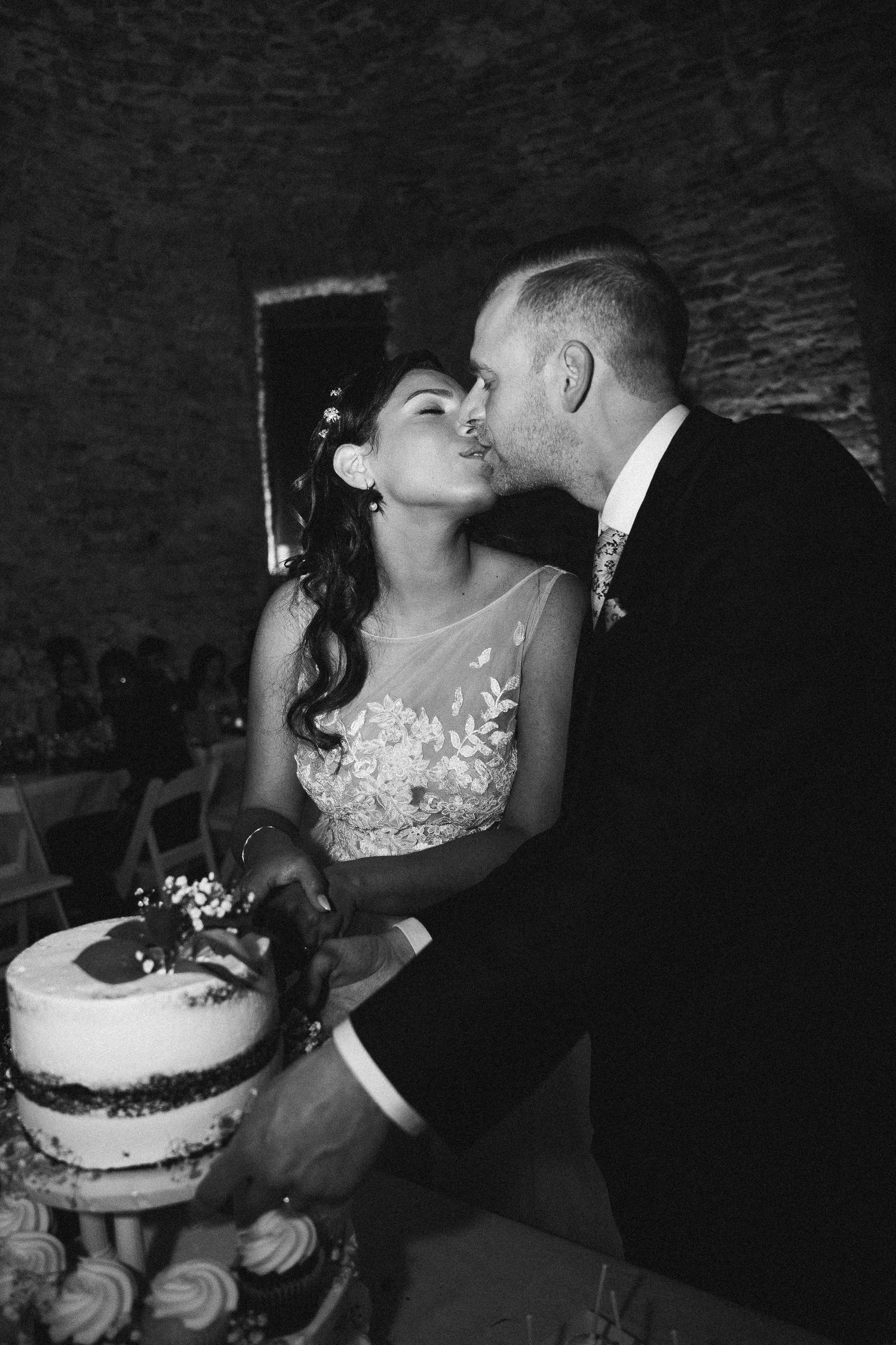 Black and white photo of newlyweds kissing while cutting their wedding cake at the reception.