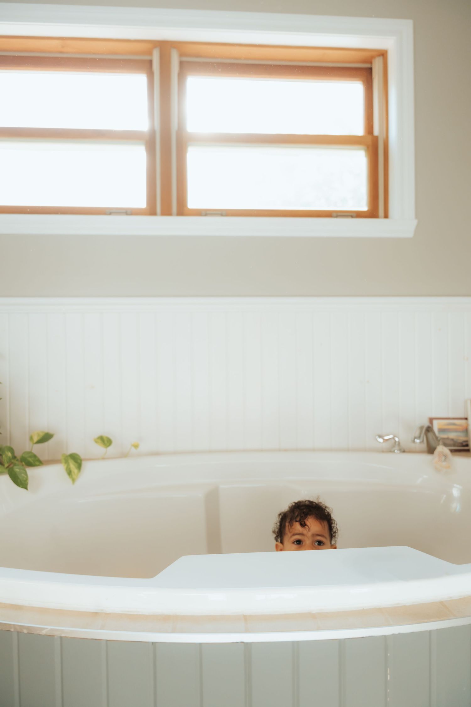 Young child peeking over the edge of a large white bathtub near a sunny window.