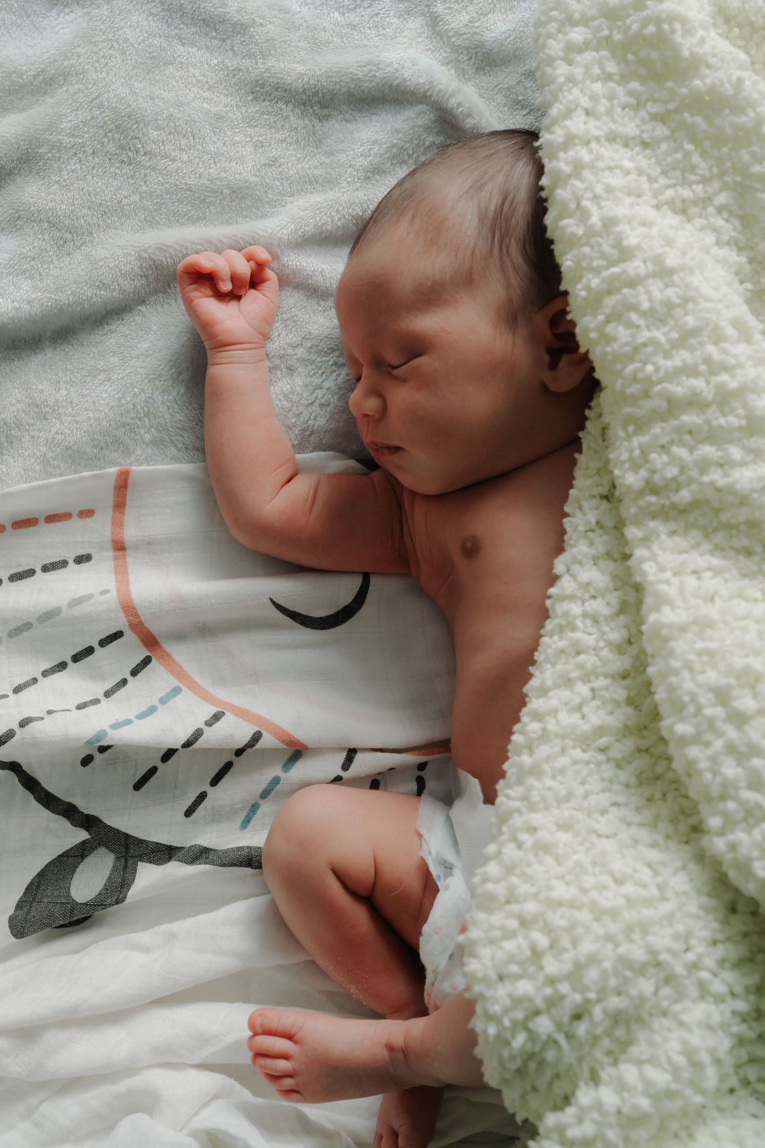 Sleeping newborn baby with fist raised lying on patterned blanket partially covered by soft white blanket.