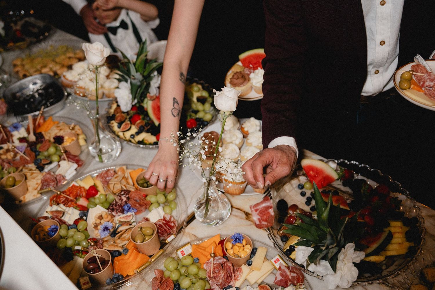 Guests selecting food from an elaborate wedding grazing table with fruits, cheeses, and charcuterie.