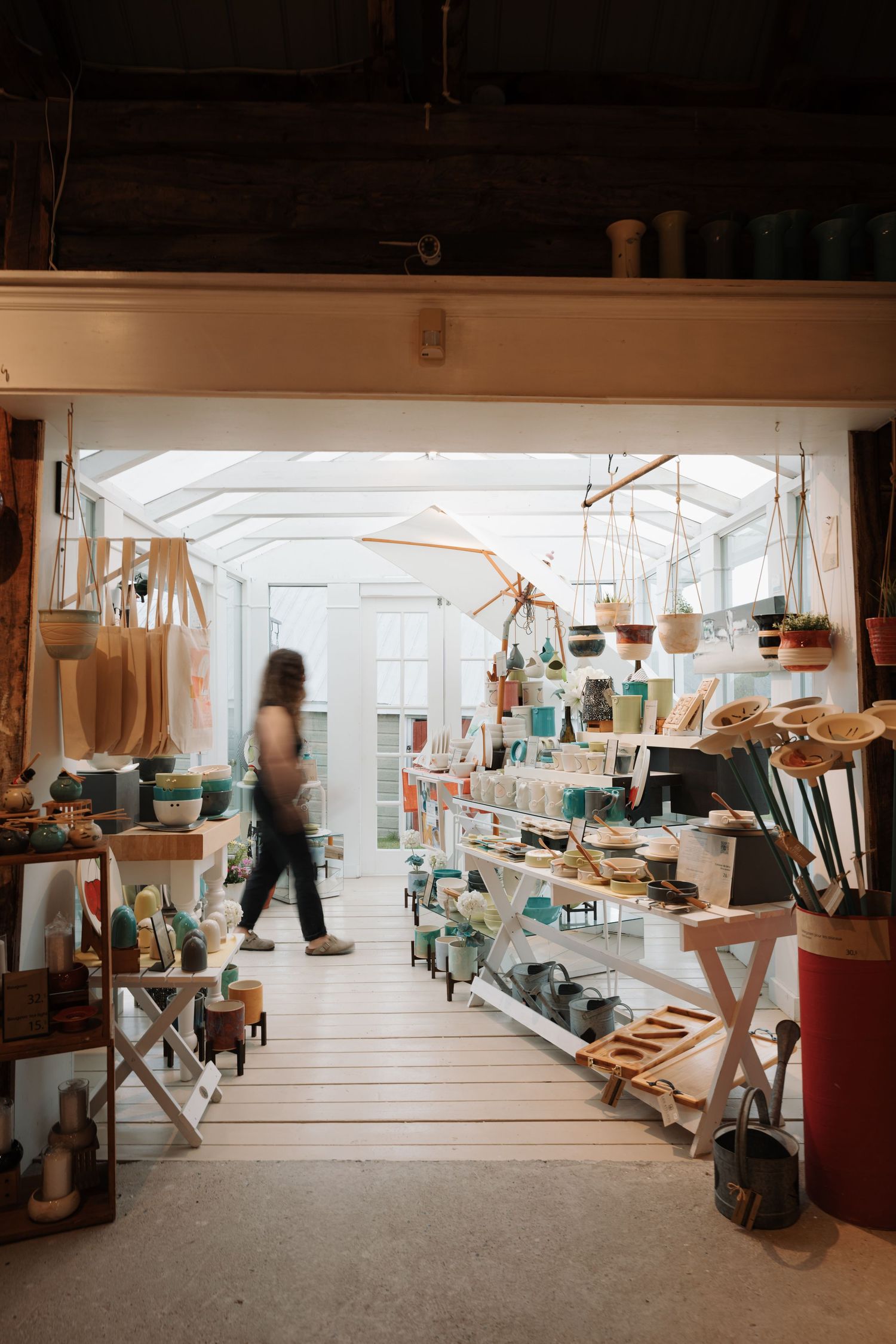 A woman walks through a bright artisan shop filled with handmade ceramics, baskets, and wooden goods displayed on shelves.