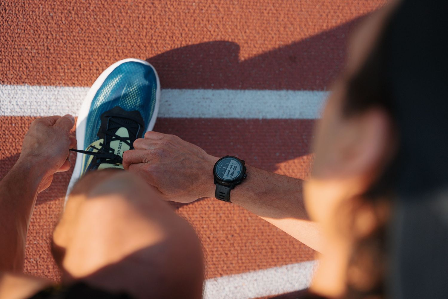 Runner tying blue sneakers on a red track with a GPS sports watch nearby.