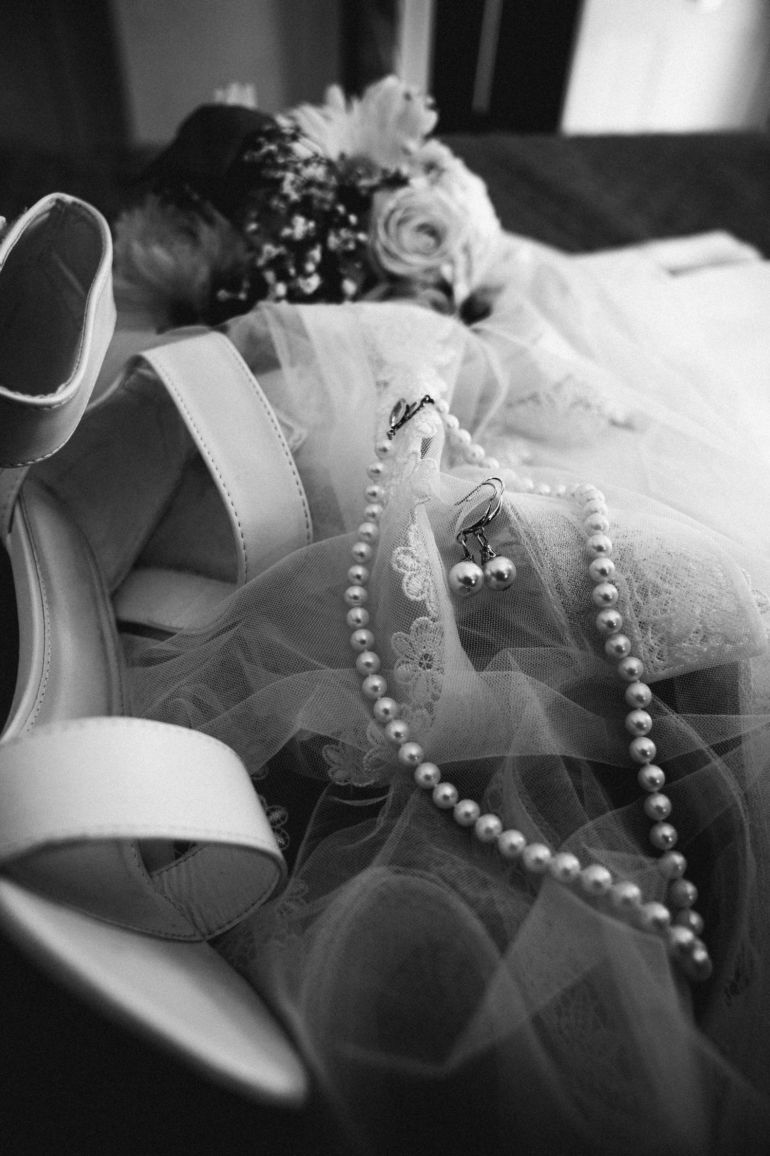 Black and white flat lay of bridal accessories including heels, pearl necklace, earrings and wedding dress.
