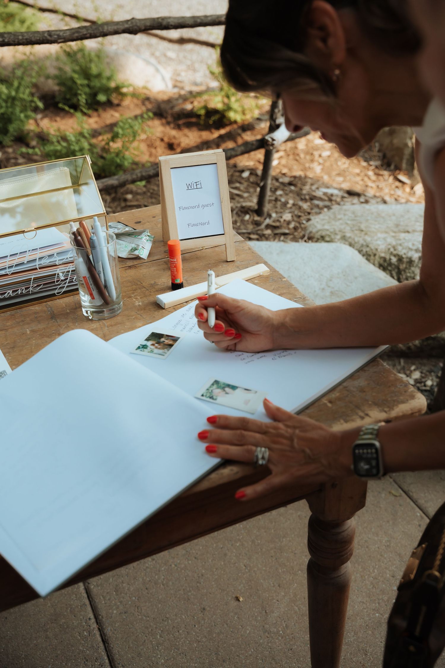 Wedding guest signing a guestbook at an outdoor wooden table decorated with photos and stationery.