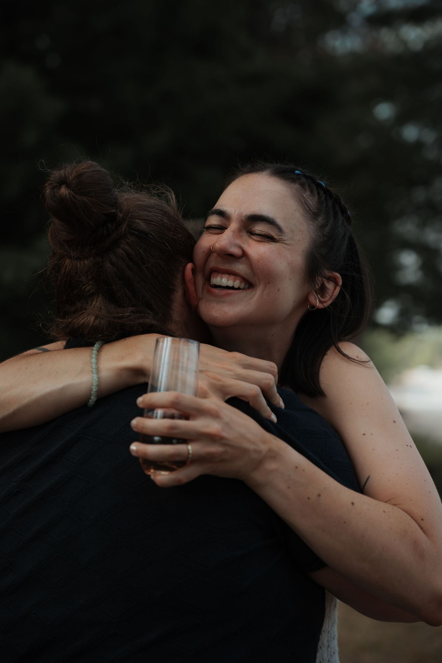 A smiling woman with dark hair joyfully hugs another guest while holding a glass at an outdoor celebration.