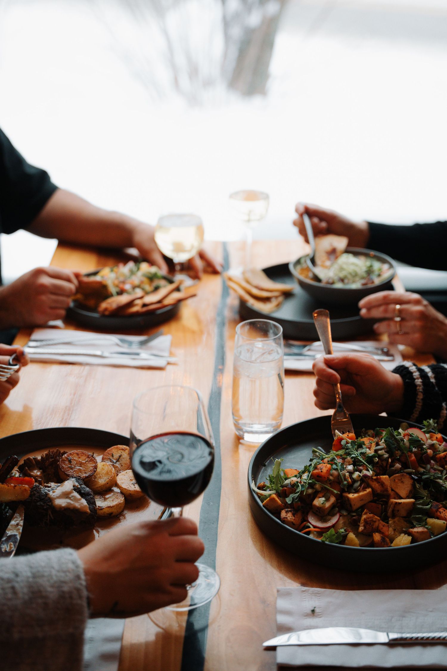 Group of diners sharing multiple dishes including salads and roasted vegetables with wine at wooden table.