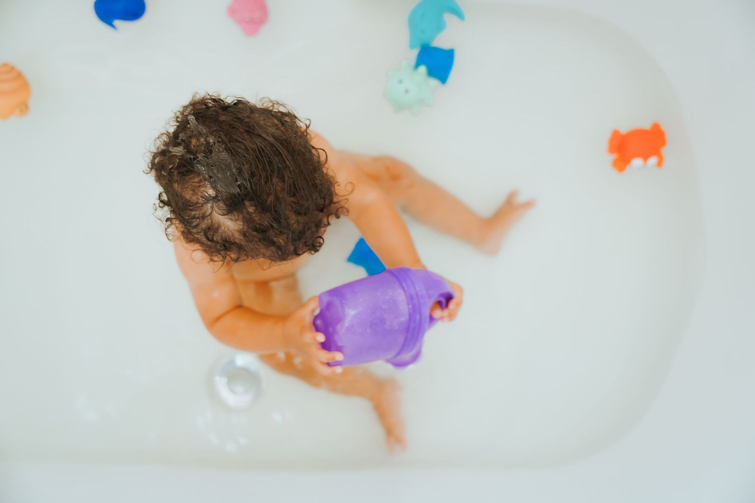 Overhead view of a young child playing with colorful bath toys in a white bathtub.