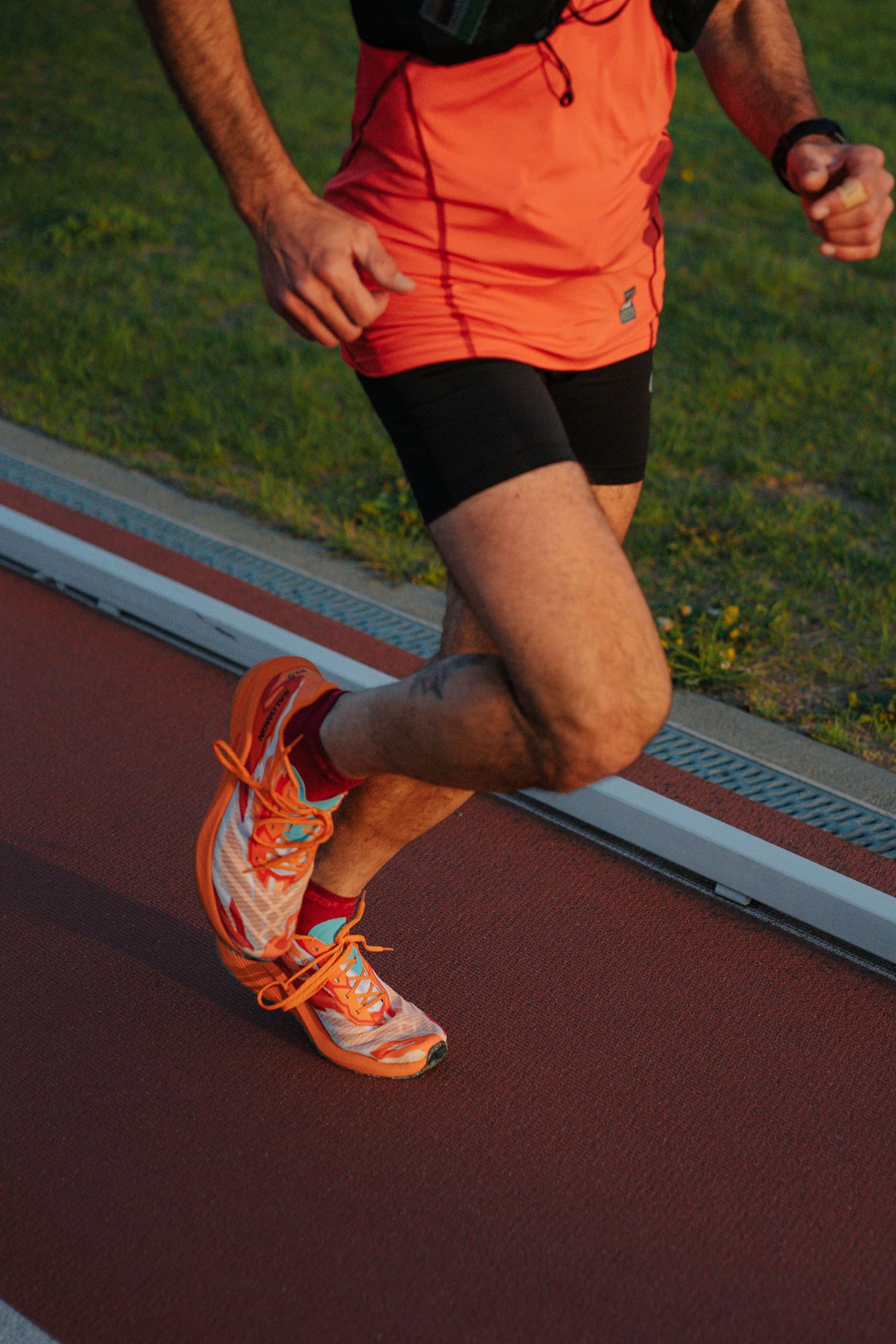 Male runner in orange vest and shorts sprinting on a red athletic track at sunset.