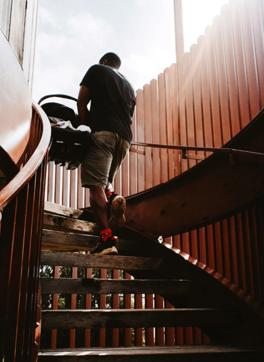 A father carries a baby car seat up wooden outdoor stairs in warm backlit sunlight.