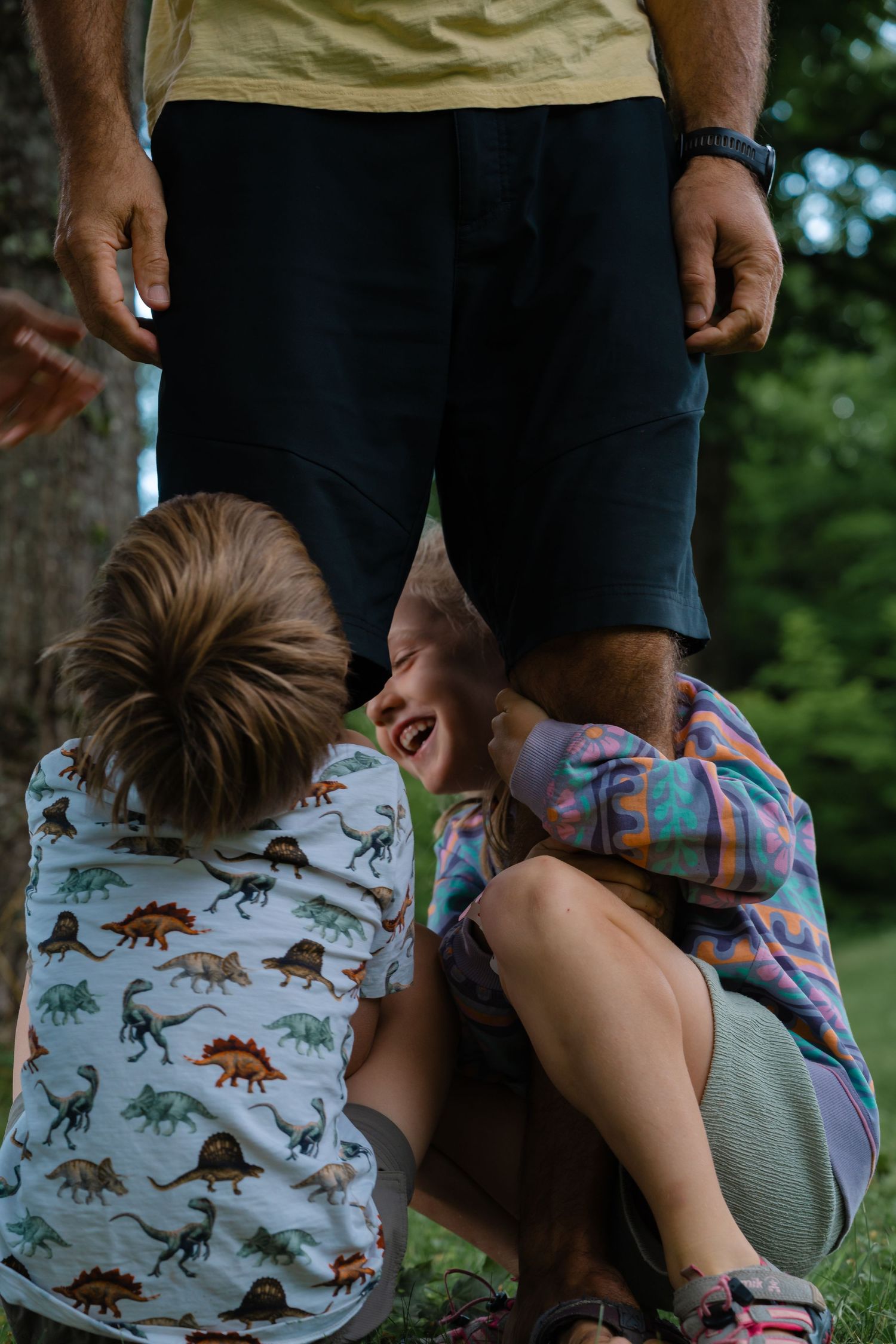 Two children laughing together outdoors near an adult in black shorts, one wearing a dinosaur print shirt.