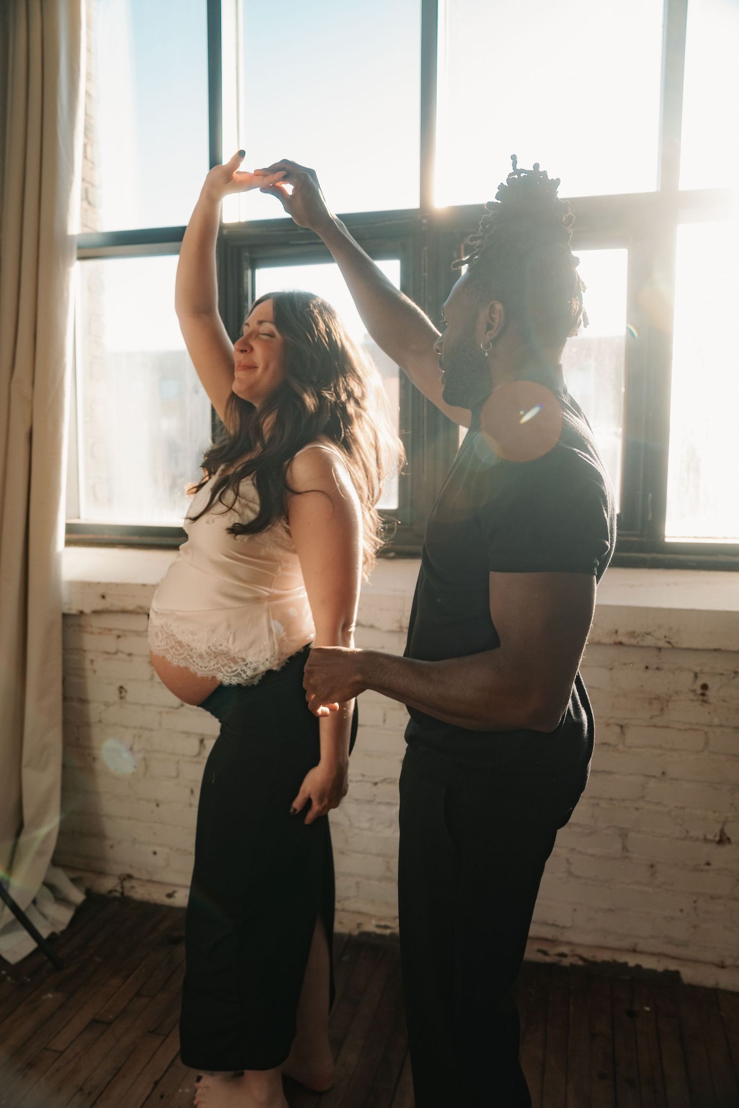 A pregnant woman dances joyfully with her partner near sunlit windows in a bright, airy room.