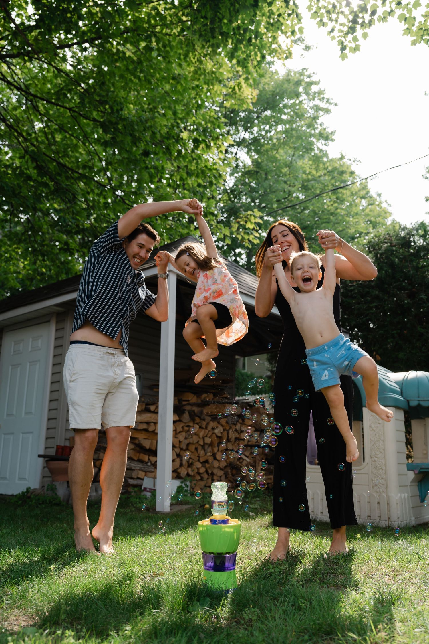 Family of four joyfully lifting children in sunny backyard with green trees and wood pile in background.