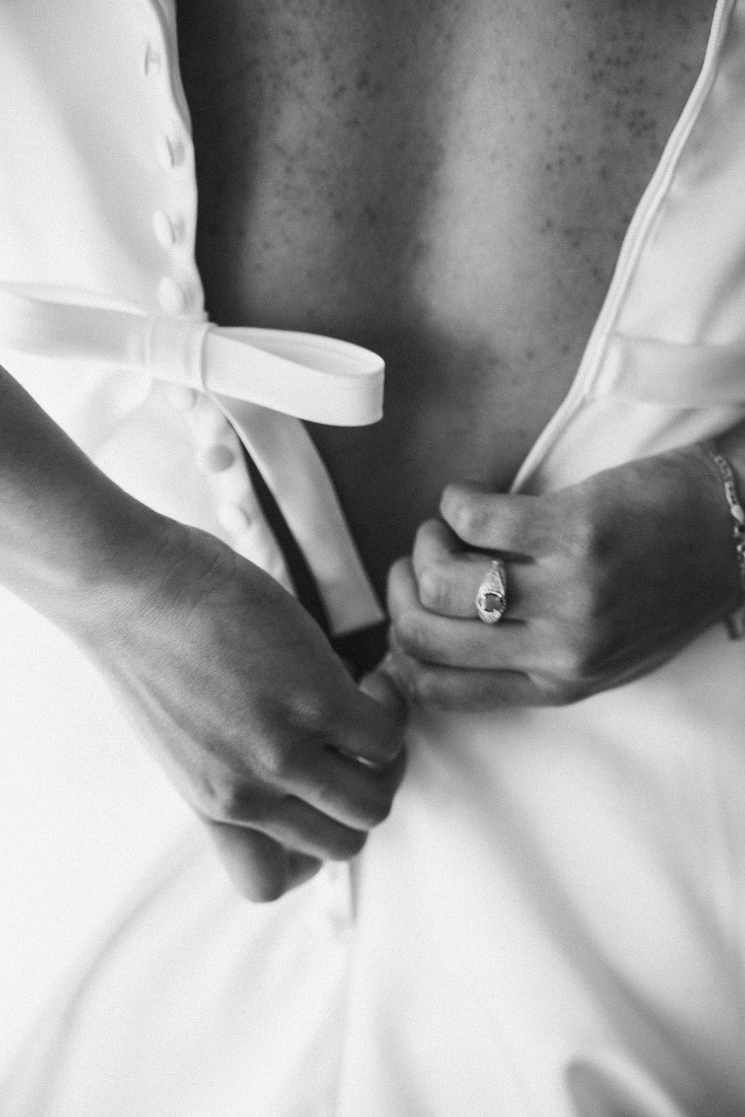 Black and white photo of a bride tying her wedding dress bow, showcasing her elegant engagement ring.