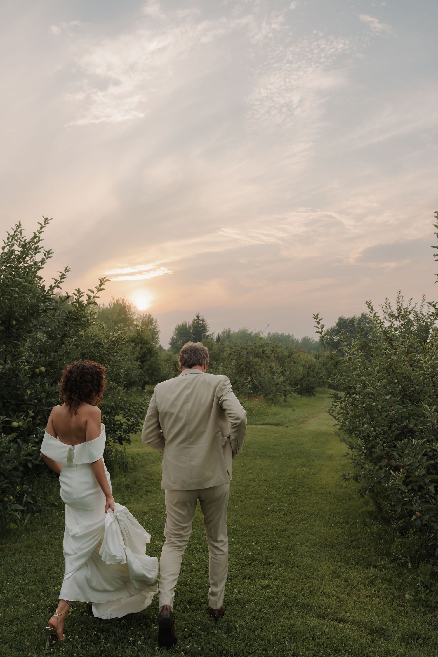 Bride and groom walk hand in hand through a lush green orchard at sunset.