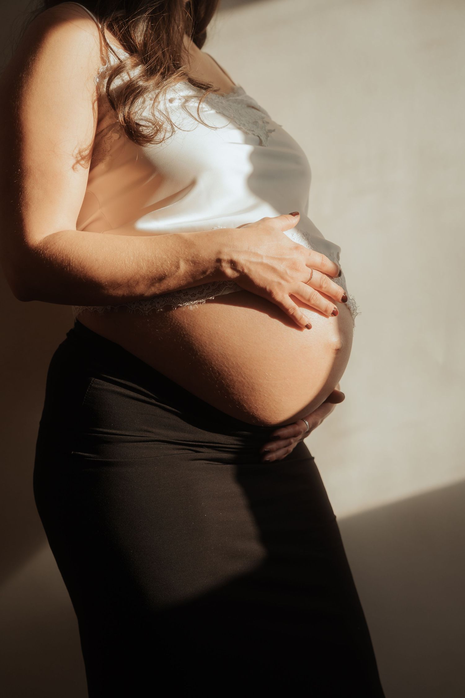 Close-up side profile of pregnant woman in white top and black skirt cradling bare baby bump in warm sunlight.