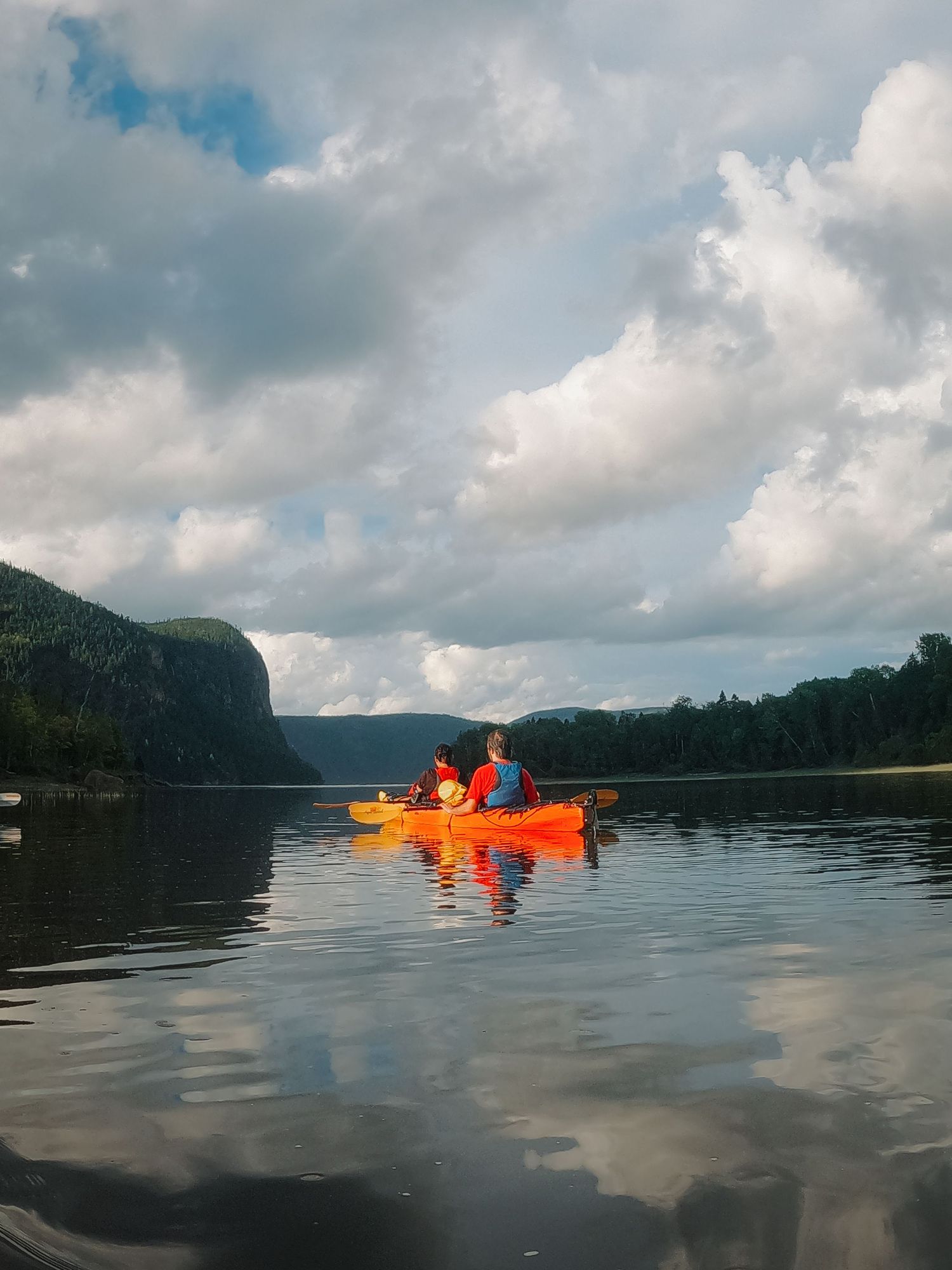 Two kayakers in an orange boat paddling across a still mountain lake under a dramatic cloudy sky.