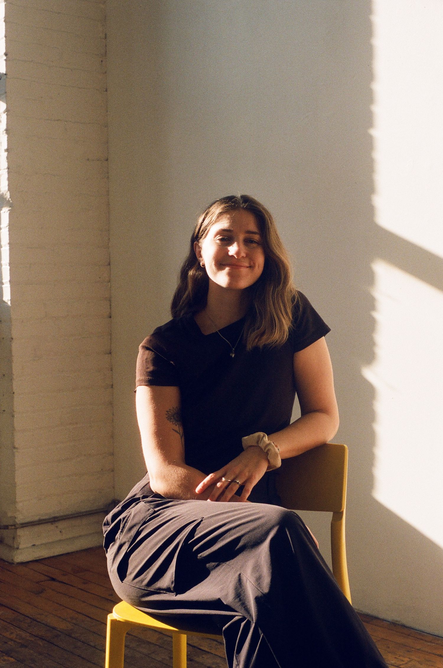 Young woman sitting on a yellow chair smiling in sunlit room with white brick wall.