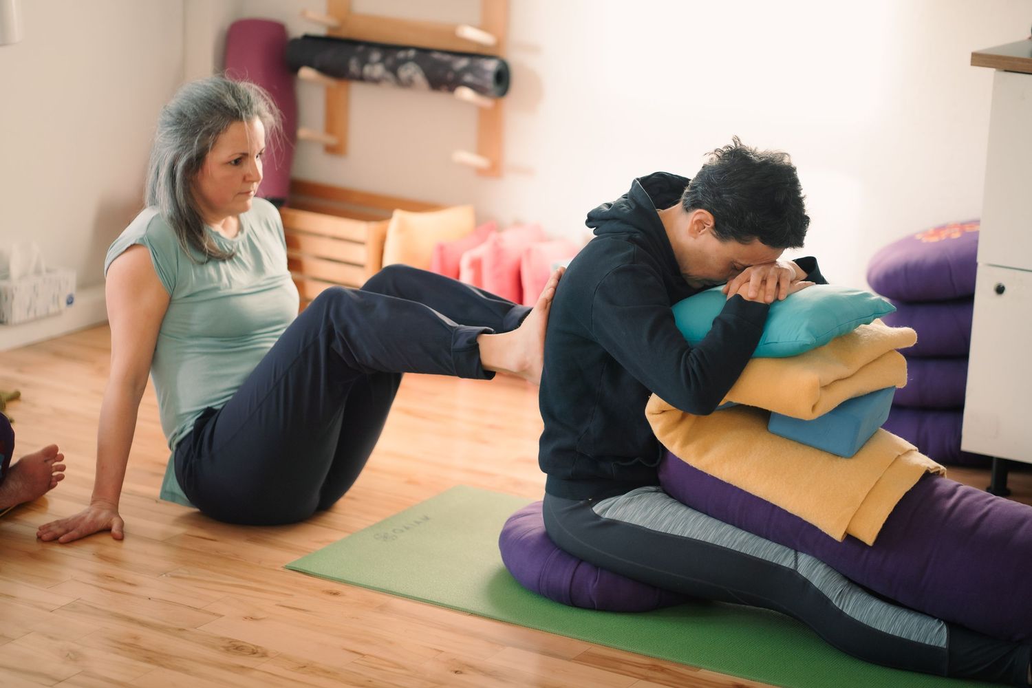 Two women practicing yoga in a bright studio, one doing sit-ups on a green mat while the other rests on cushions.