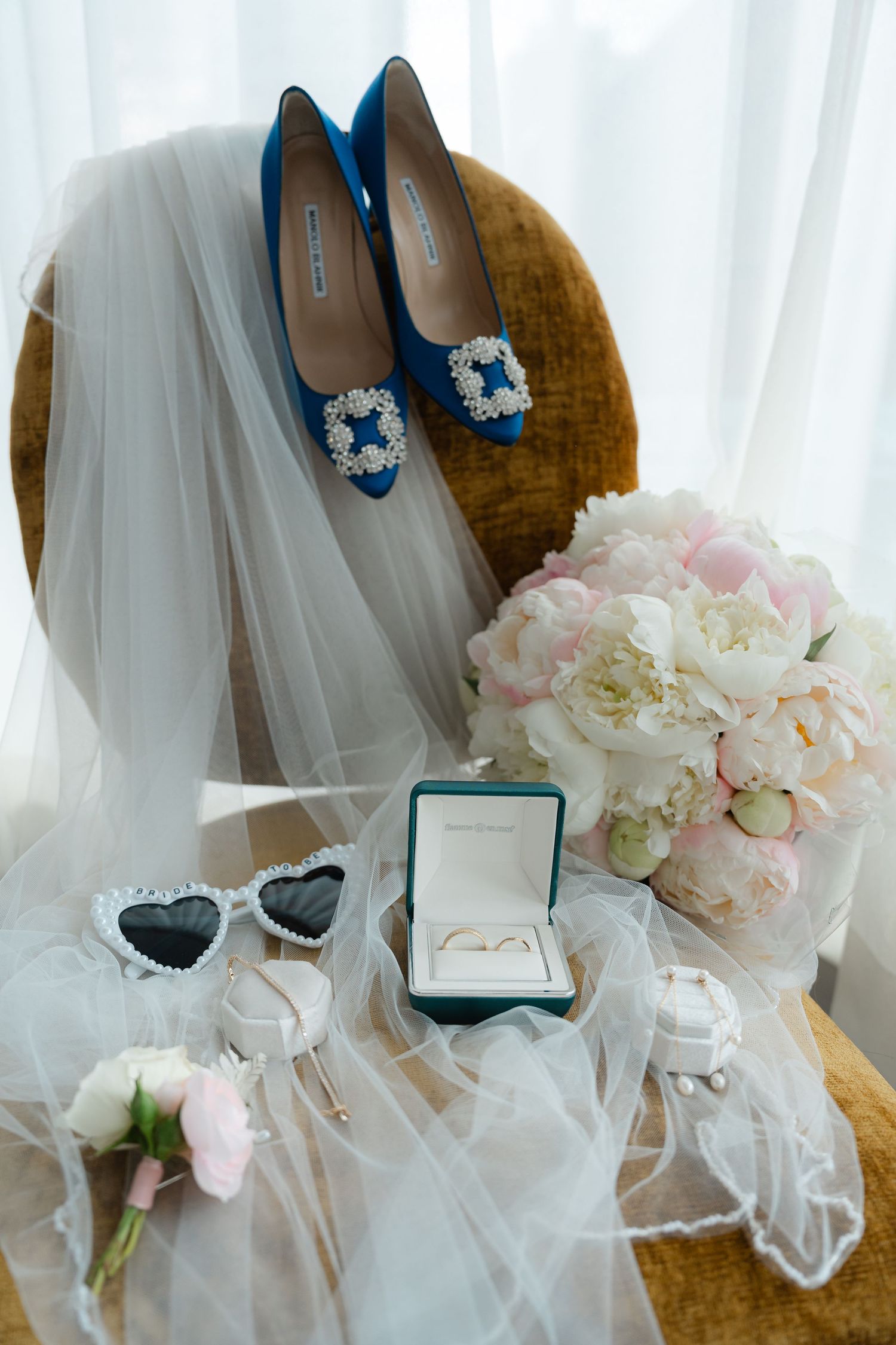 Blue bridal heels with pearl buckles, wedding rings in teal box, heart sunglasses, bouquet, and veil arranged on velvet chair.