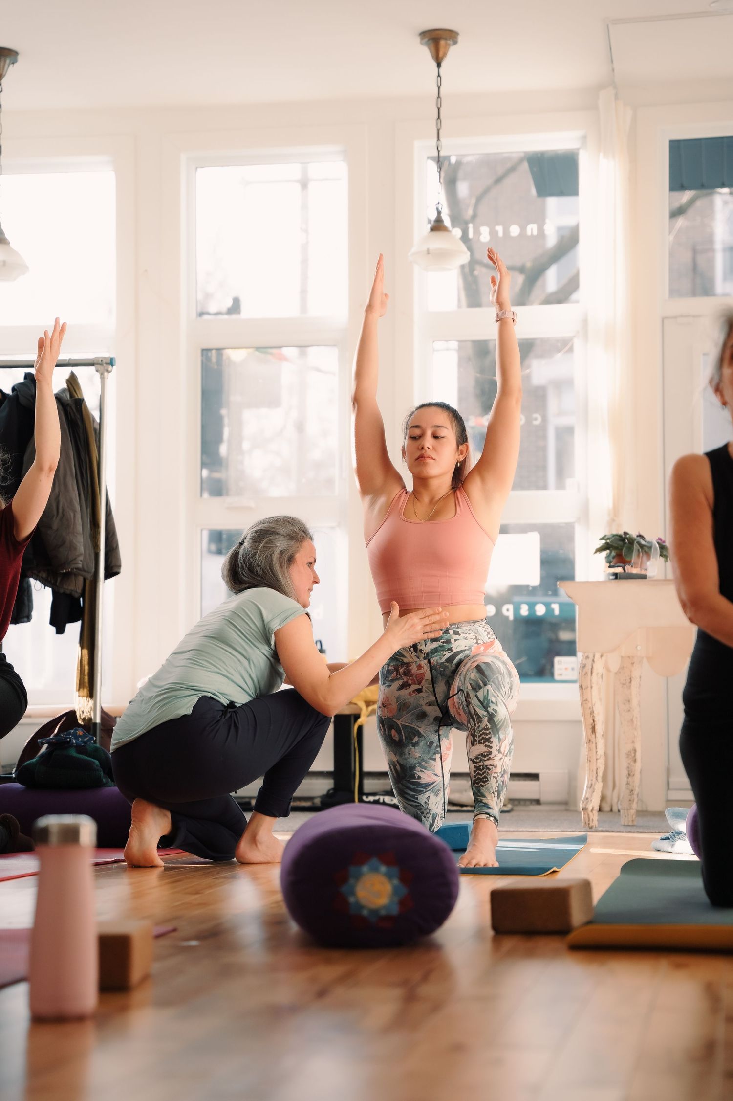 Yoga instructor assisting student in a low lunge pose with arms raised in a sunlit yoga studio.