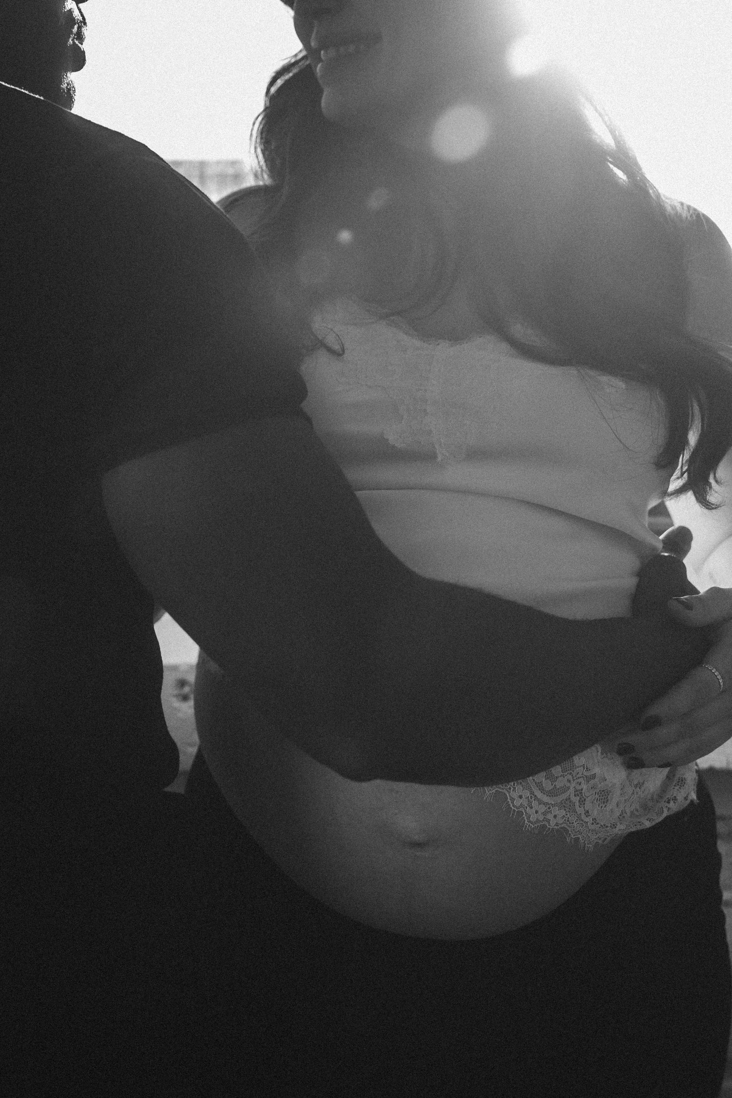 Black and white backlit silhouette of pregnant woman being embraced by partner in sunlight.