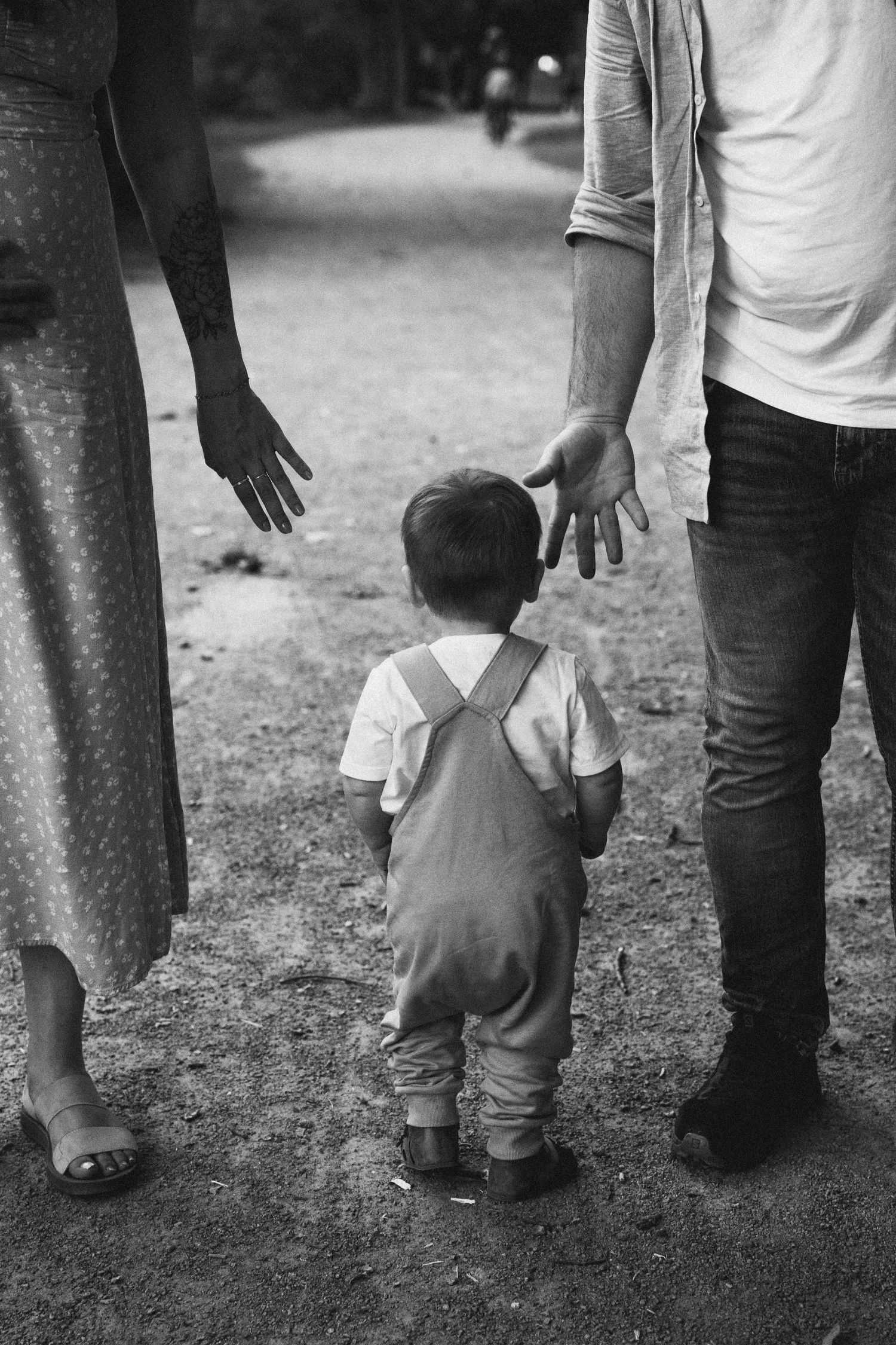 Black and white photo of a toddler walking between two parents on a path.