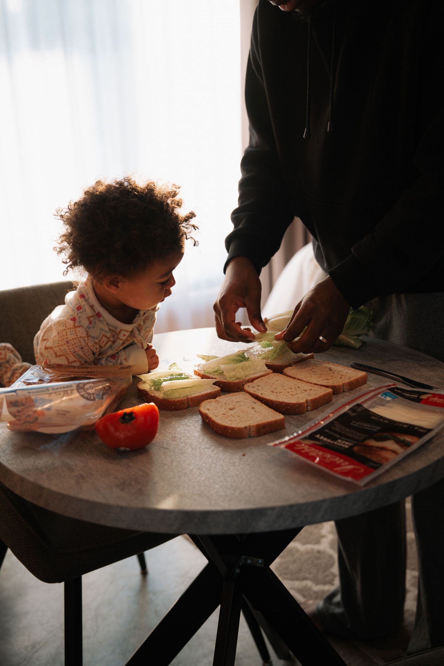Curly-haired toddler watching adult prepare sandwiches with vegetables on round table.
